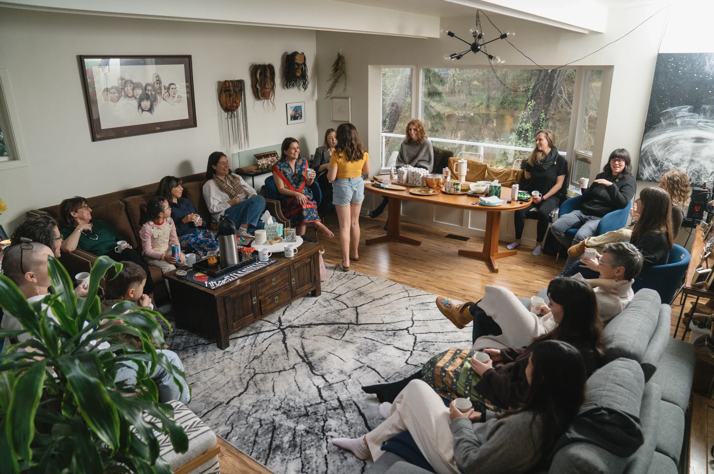 Group of people gathered in a cozy living room, sitting on sofas and chairs, with a table of snacks and drinks, celebrating in a relaxed, informal setting.