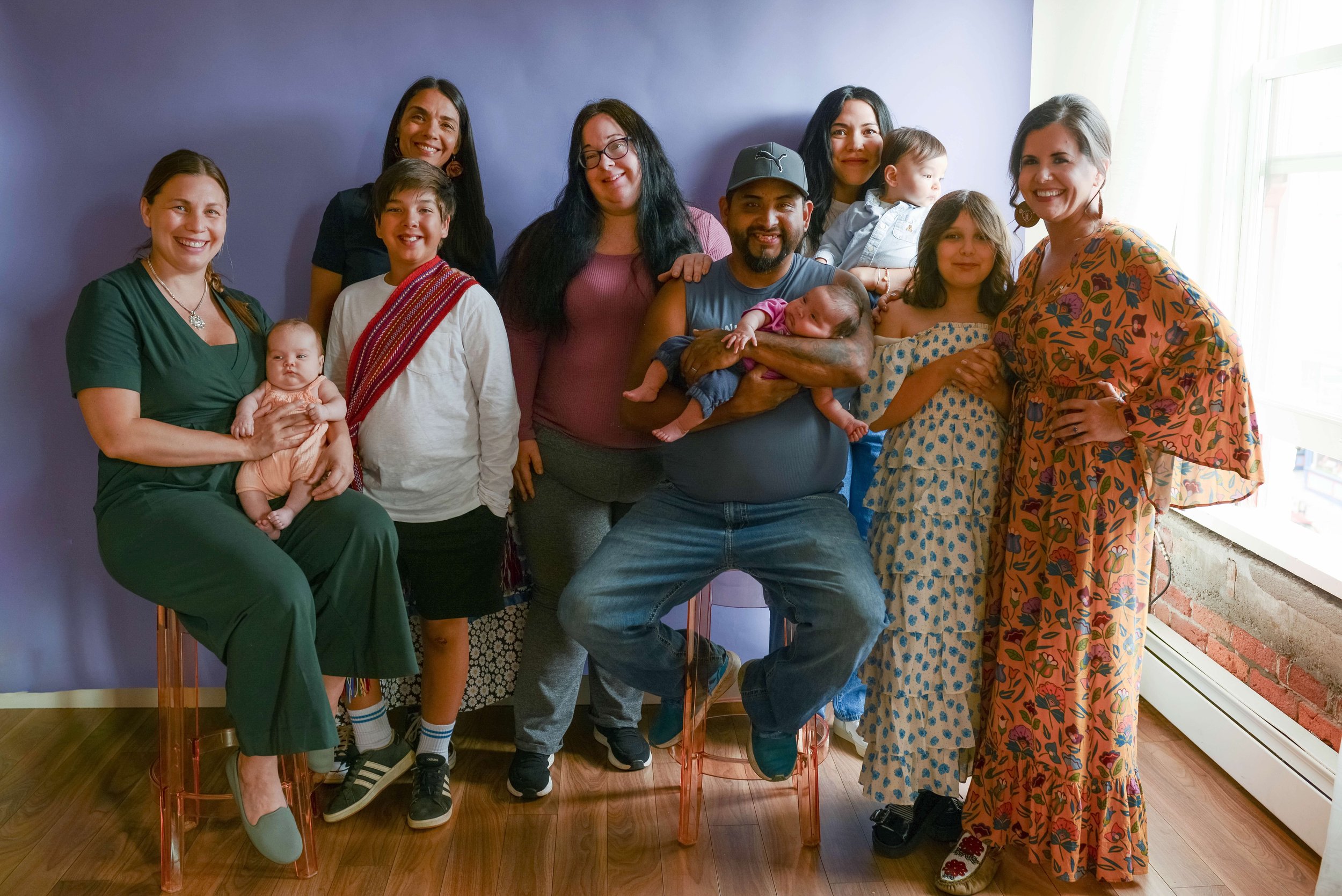 Group of people, including adults and children, posing together indoors near a purple wall and a window with curtains. Several individuals are smiling, and one person is holding a baby.