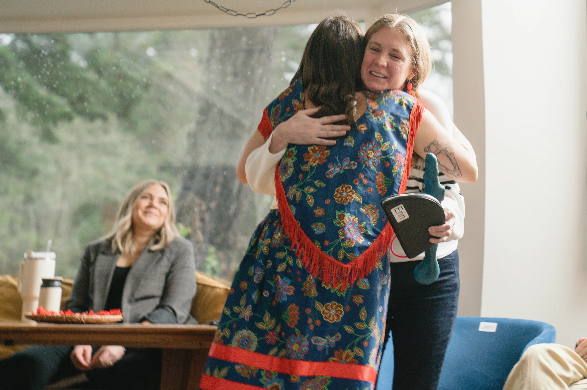 Two women are hugging each other, smiling, with a third woman seated at a table watching them and smiling. The woman on the right is holding a black case and a teal object, possibly a prosthetic or an artistic item. The scene appears to be in a cozy indoor space with a large window showing a green outdoor background.