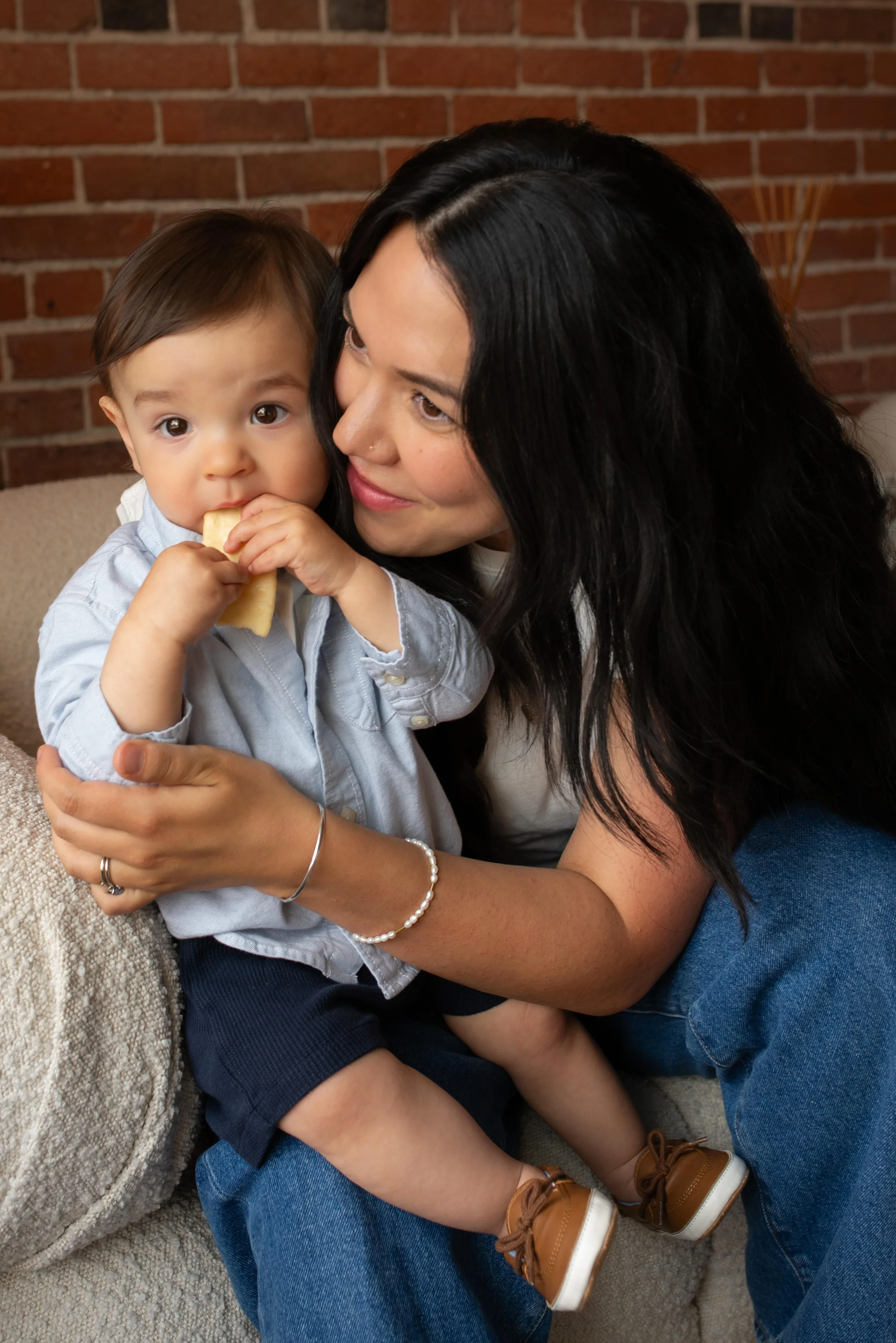 A woman with long black hair holding a young boy with short brown hair, who is eating a cracker, sitting on her lap indoors with a brick wall in the background.
