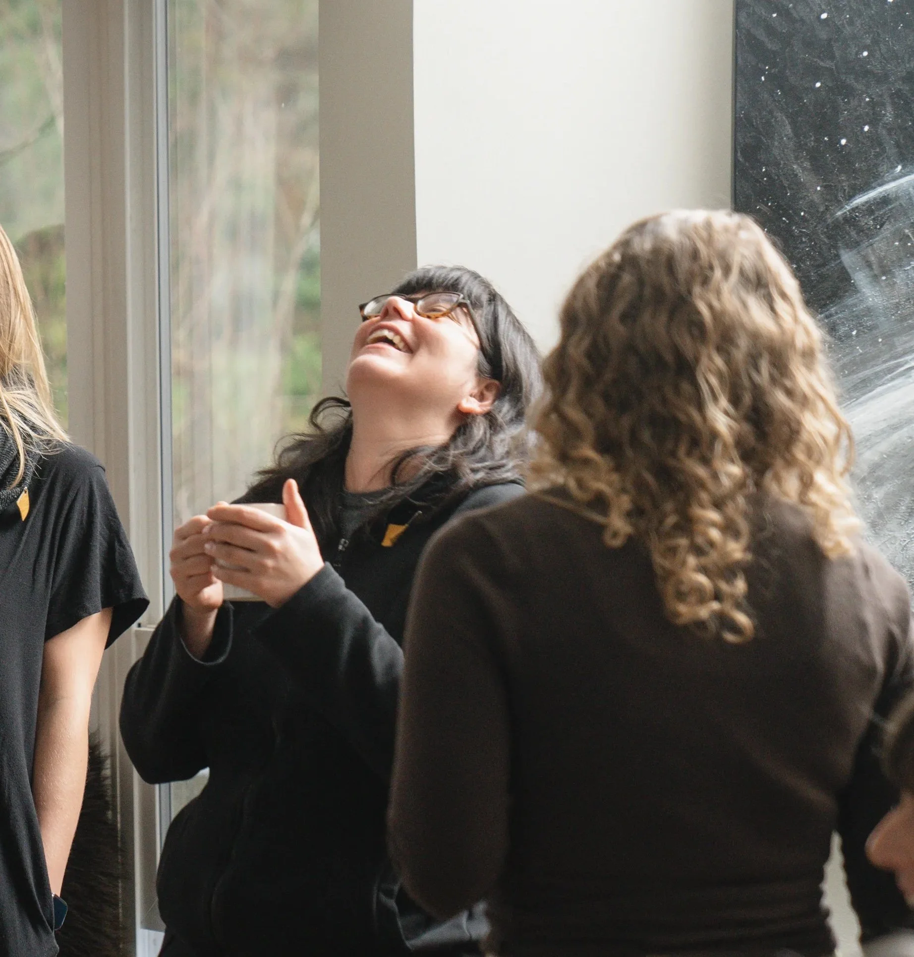 A young man sitting on a beige cushioned bench against a neutral wall, wearing a gray polo shirt and cream-colored pants, with a contemplative expression and shadow cast behind him.