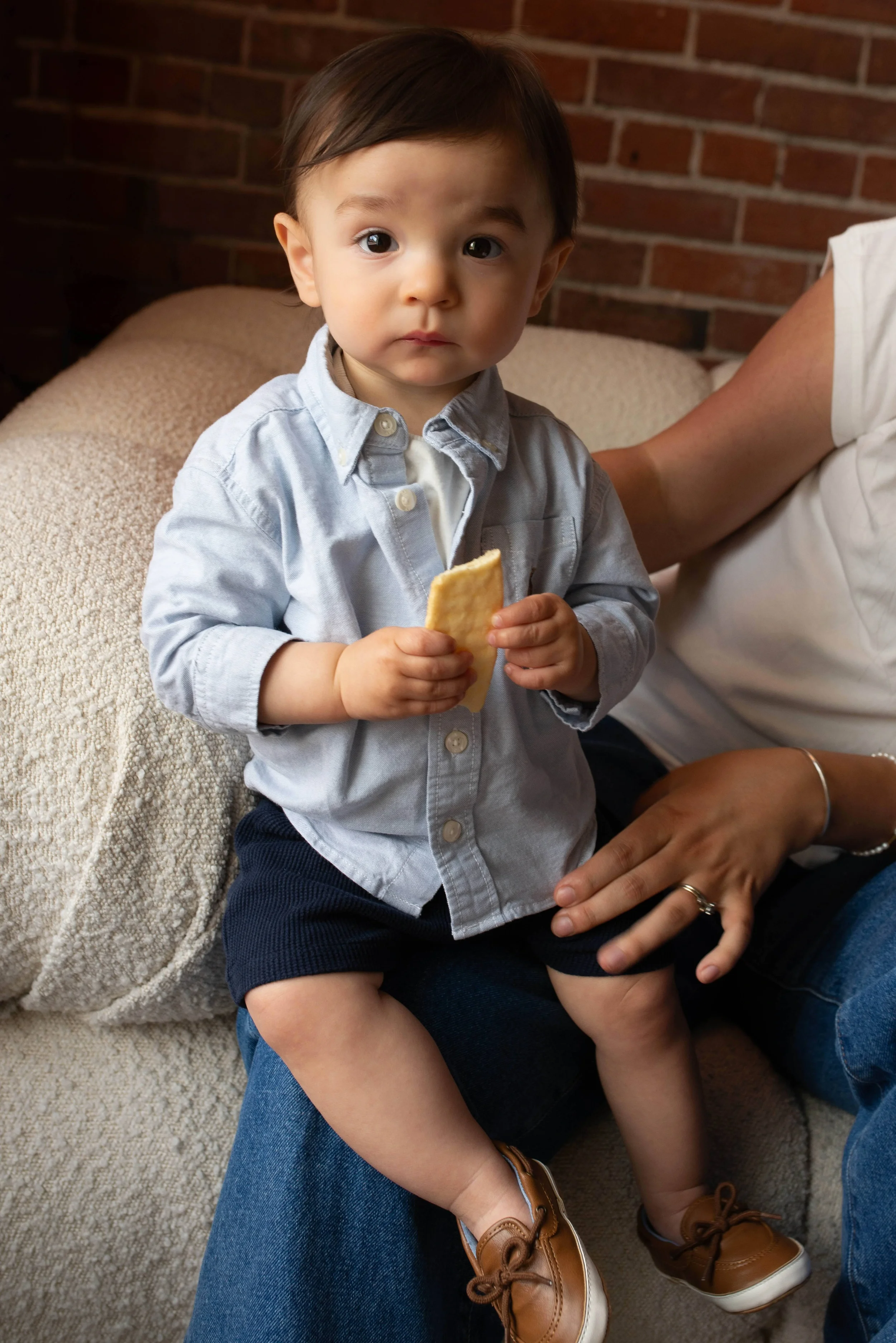 A young boy sitting on an adult's lap holding a biscuit, wearing a light gray button-up shirt, navy shorts, and tan shoes, with a brick wall in the background.