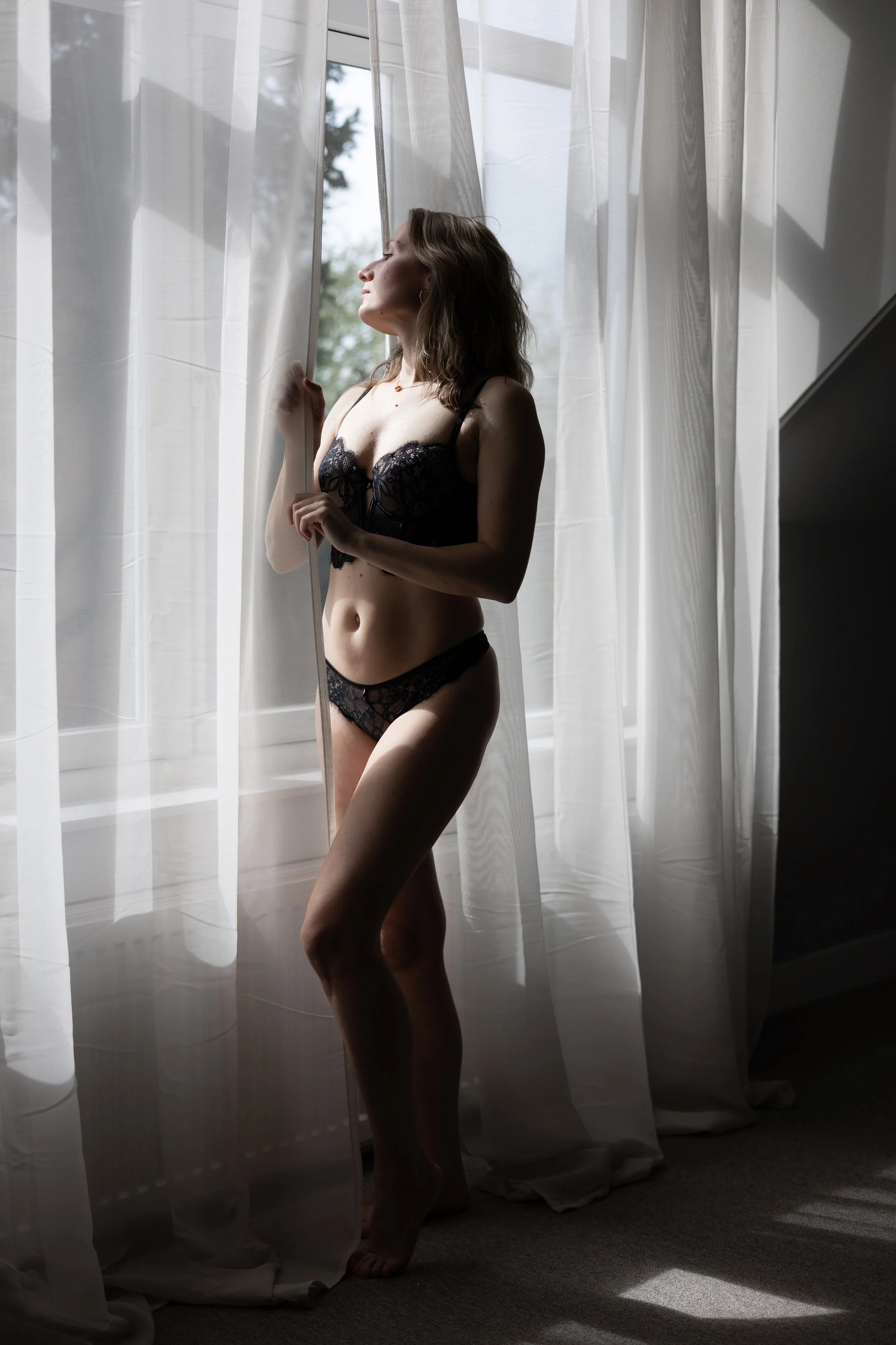 Vrouw in zwarte lingerie die achter een witte vitrage door een raam naar buiten kijkt.
