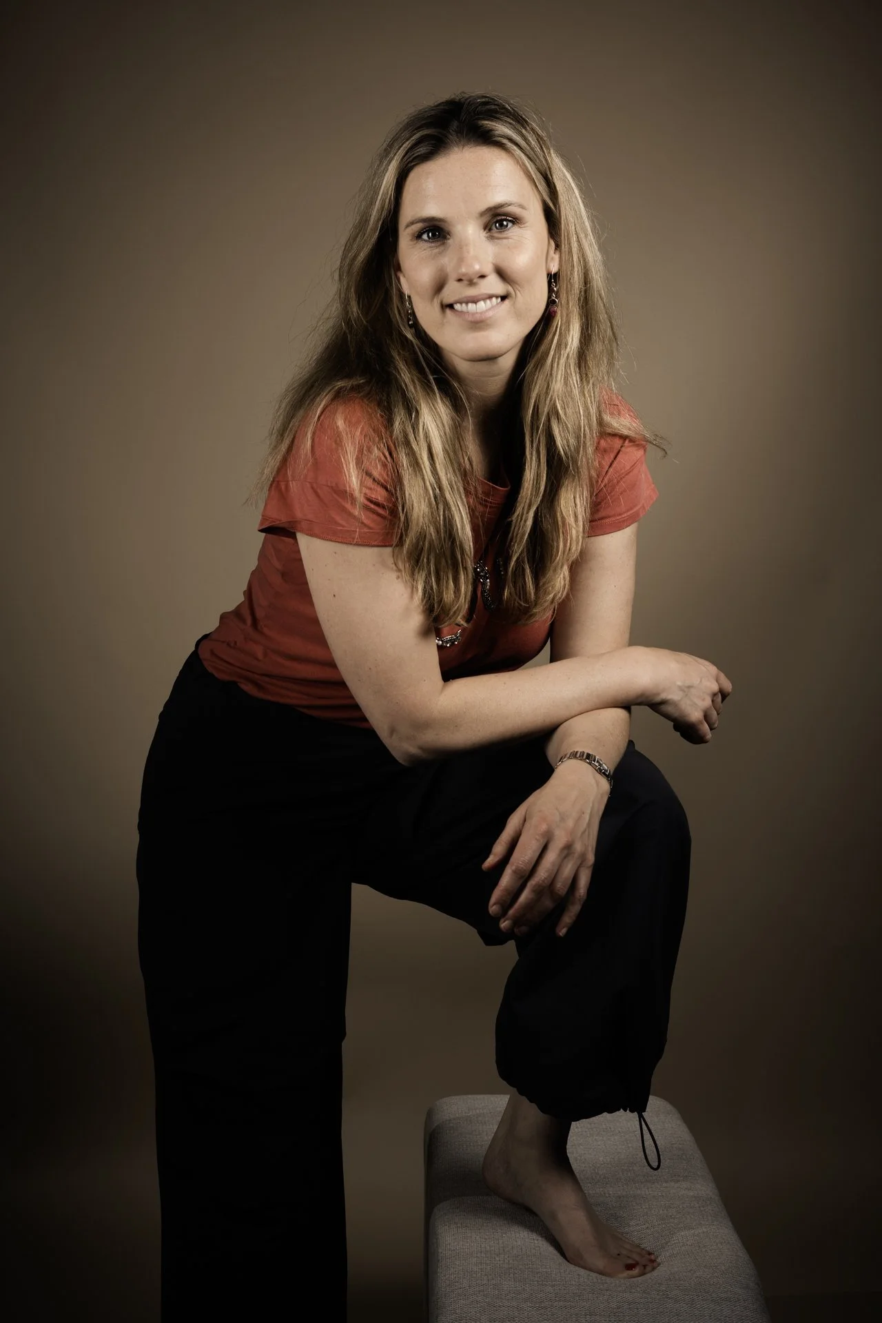 Een vrouw in een rode top en zwarte broek staat krachtig met één voet op een beige voetbank, lachend en naar de camera kijkend tegen een effen bruine achtergrond.