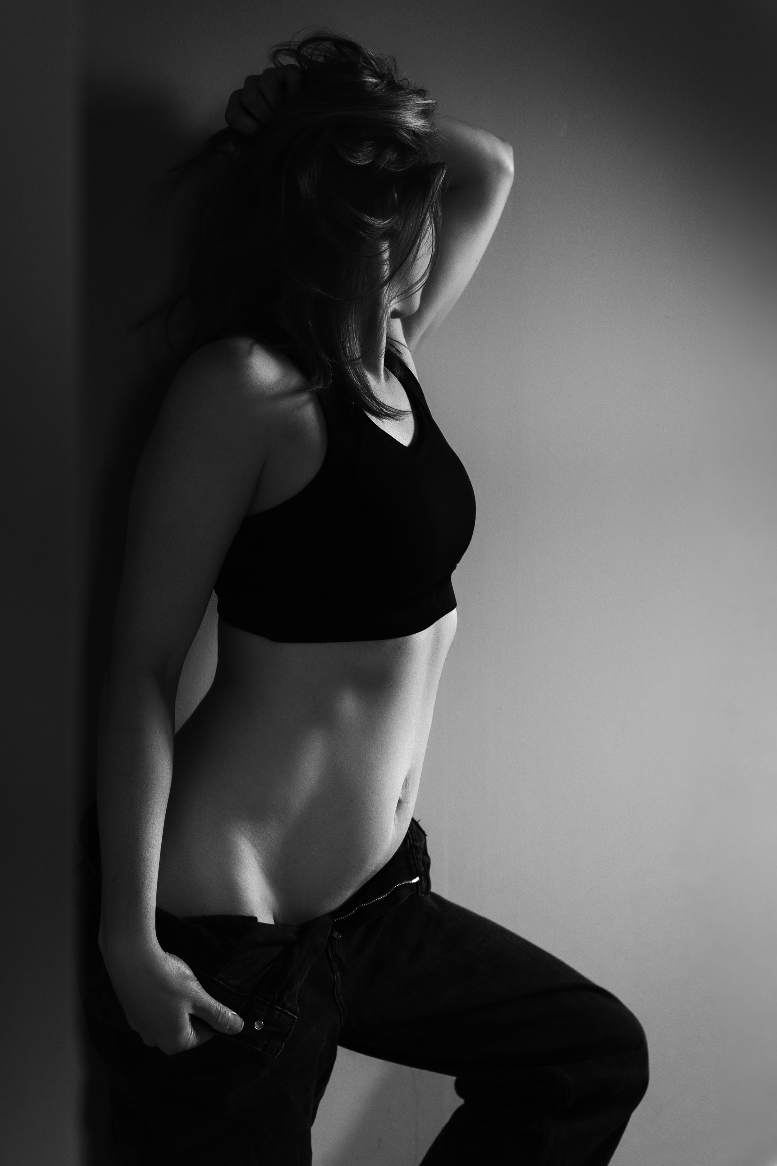 Zwart-wit foto van een vrouw die tegen een muur leunt, met één arm omhoog achter haar hoofd en de andere hand in haar broekzak, gedragen in een sportbeha en jeans.