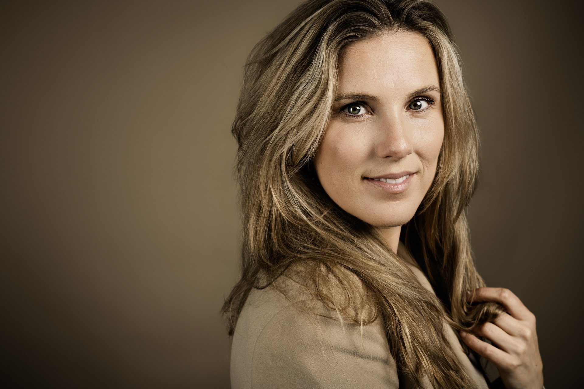 Portret van een vrouw met lang, golvend, lichtbruin haar en lichte huid, die glimlacht en recht naar voren kijkt, tegen een neutrale achtergrond.