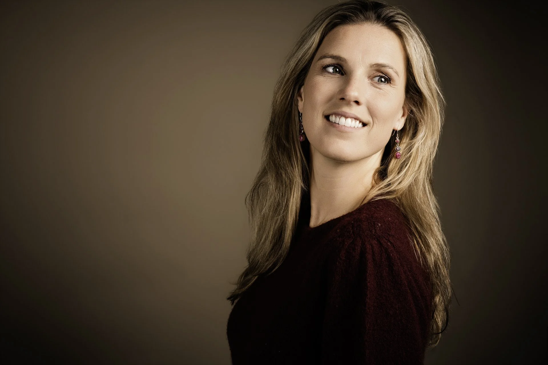 Portret van een lachende jonge vrouw met lang, golvend lichtbruin haar en oorbellen, gekleed in een donkerrode trui, tegen een effen achtergrond.