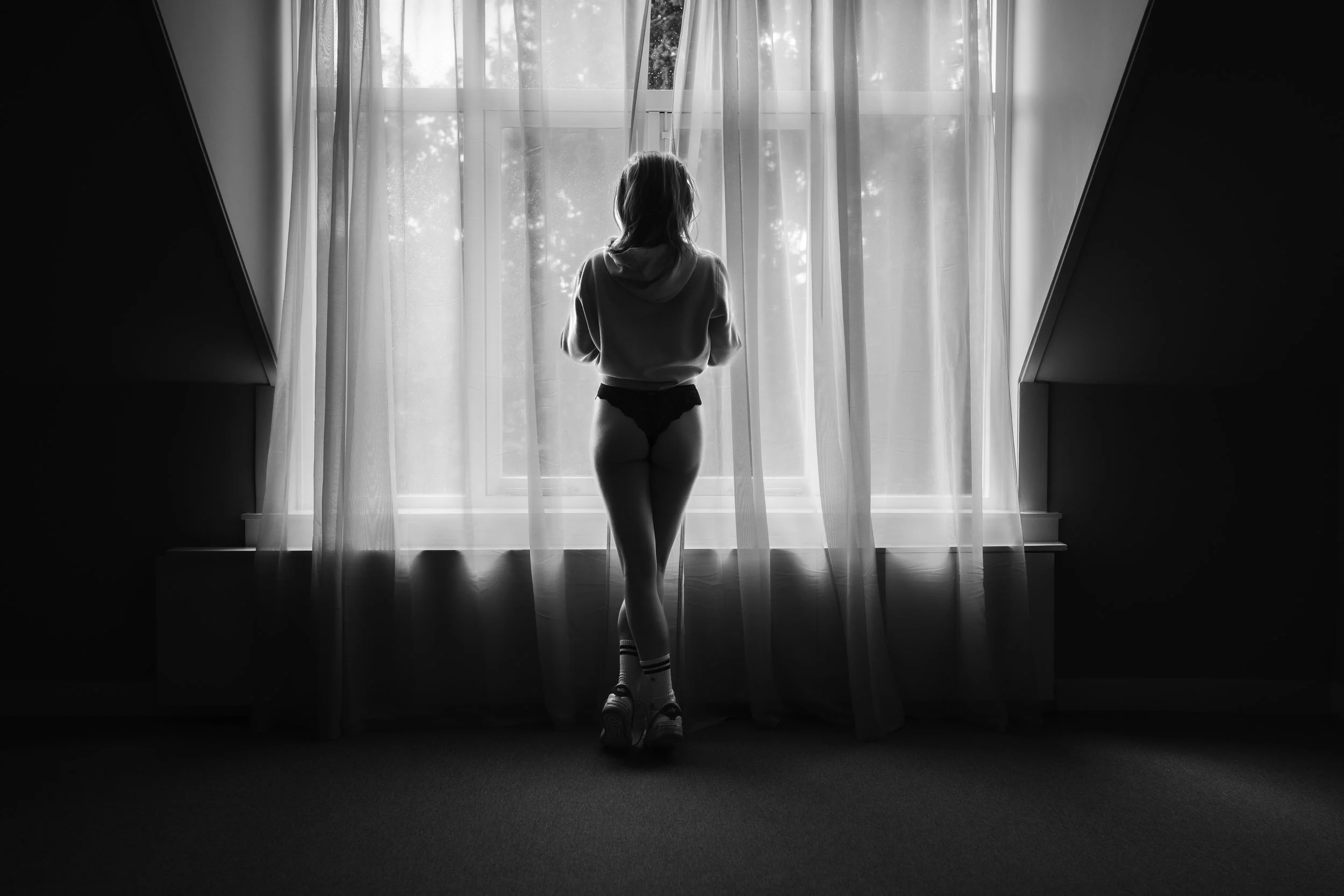 In de foto is een persoon te zien die naar een raam staart, gekleed in een hoodie en onderbroek, met sokken en schoenen. Het is een zwart-wit foto en de persoon staat in een kamer met gordijnen die het raam bedekken. Het licht van buiten schemert naar binnen.