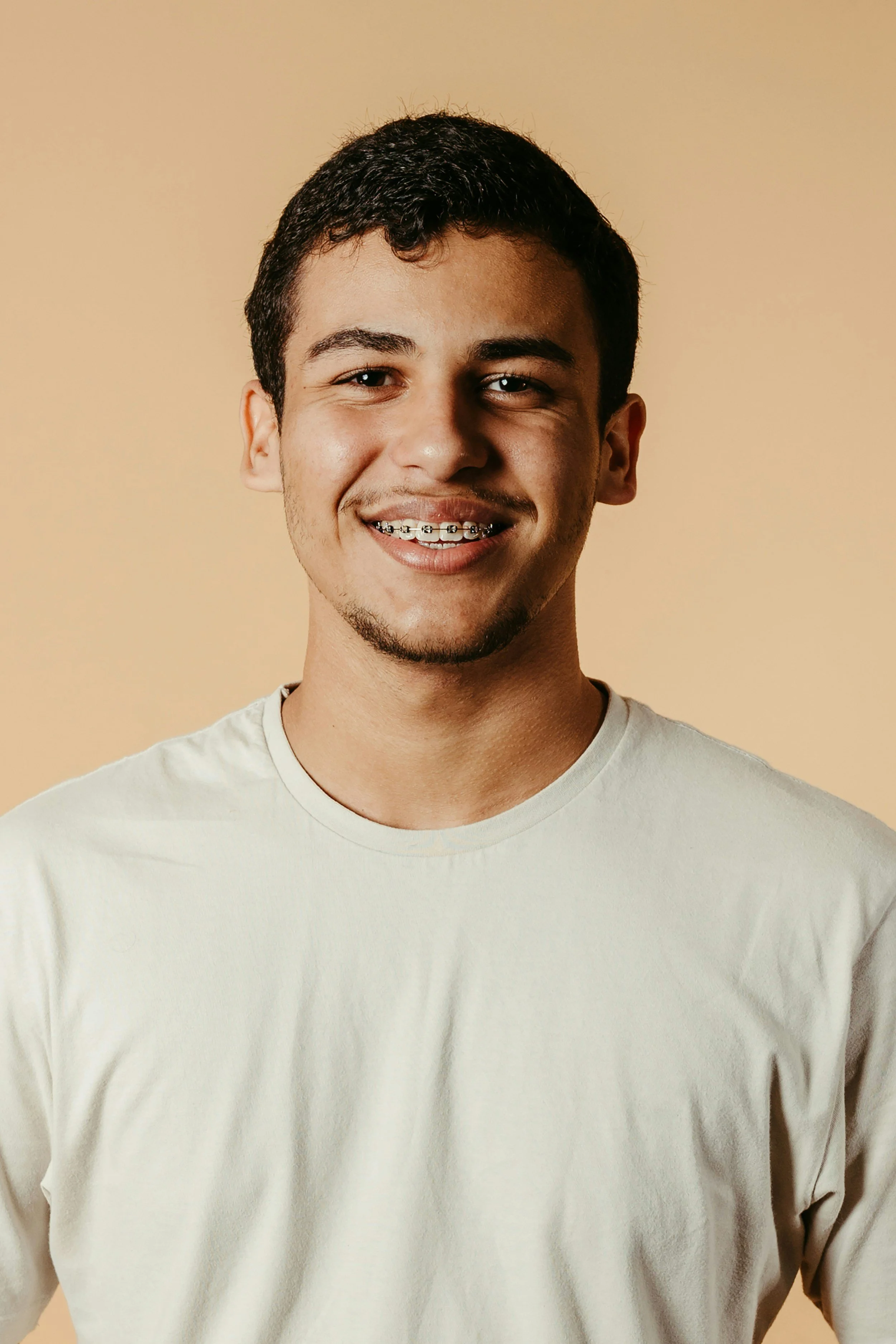A young man with short, dark hair and braces on his teeth, wearing a white t-shirt, smiling against a beige background.