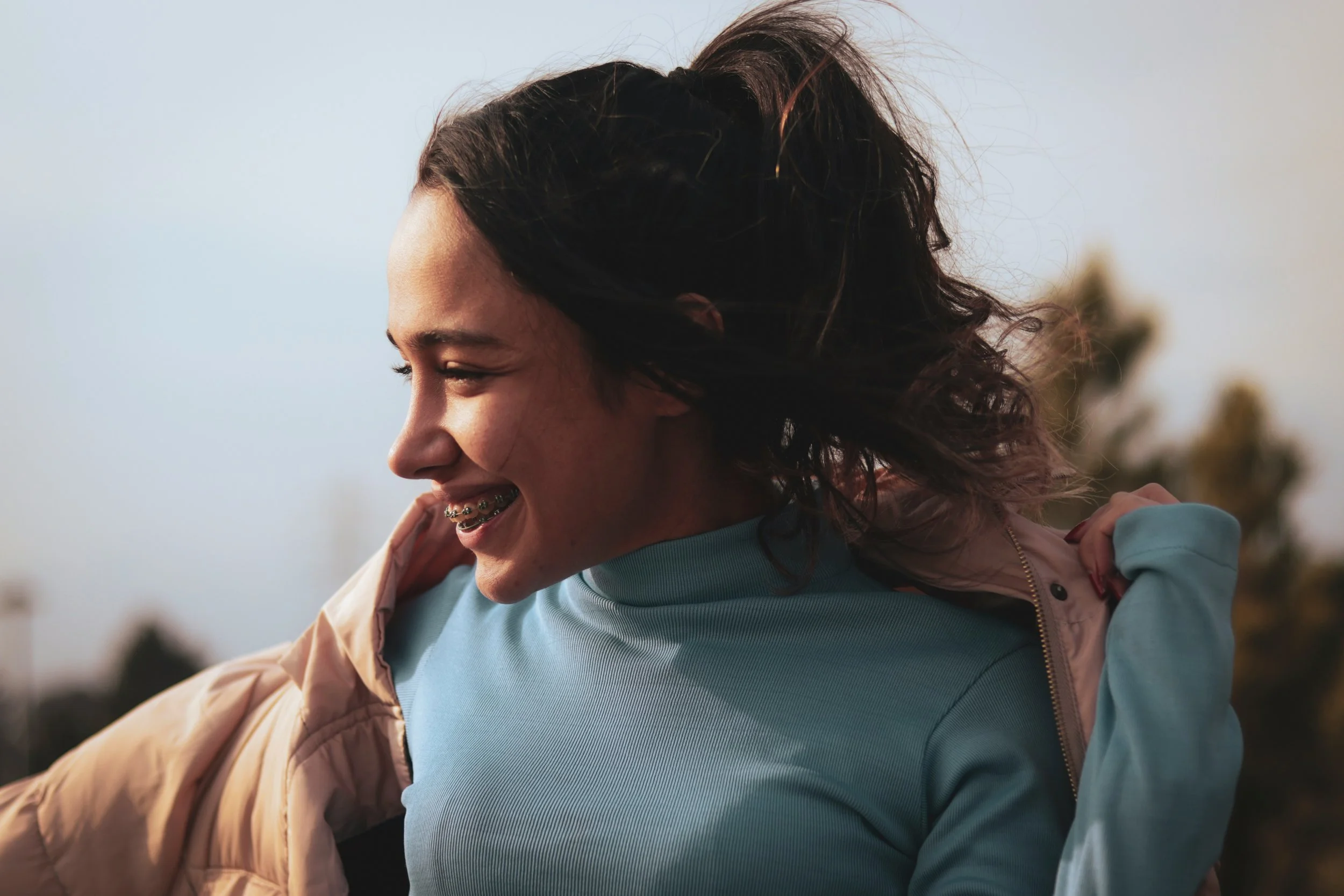 A woman smiling outdoors with wind blowing her dark hair. She is wearing a beige jacket over a light blue shirt.