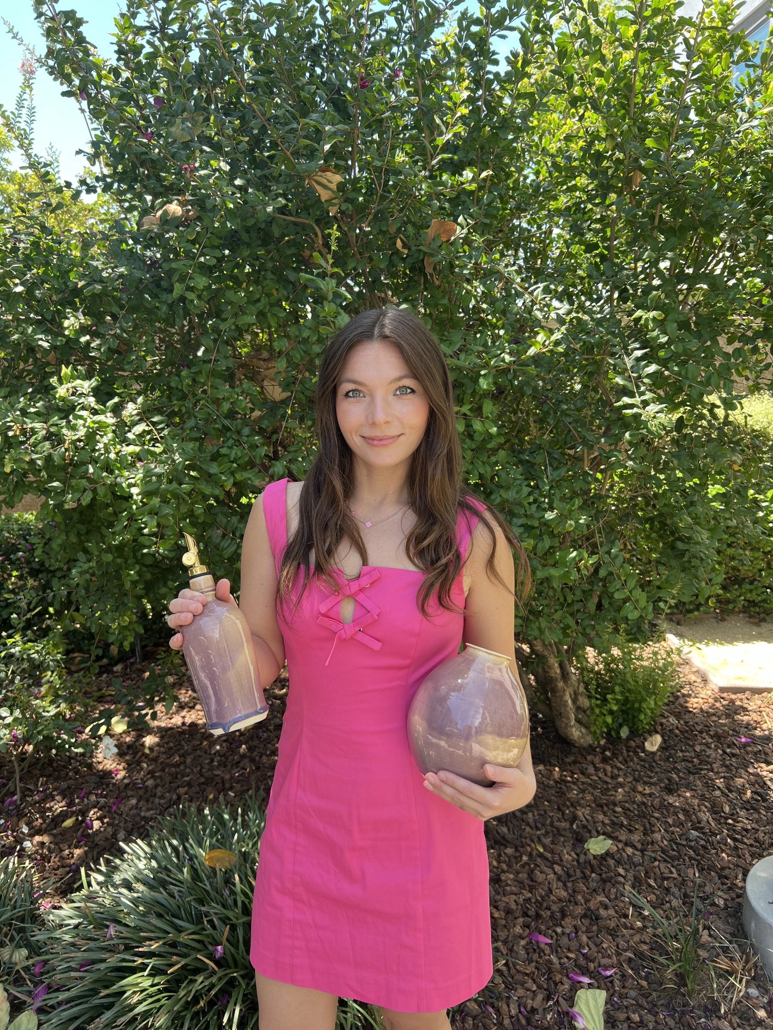 A young woman with long brown hair wearing a pink dress, standing outdoors in front of a large leafy green bush, holding a ceramic vase and olive oil bottle.