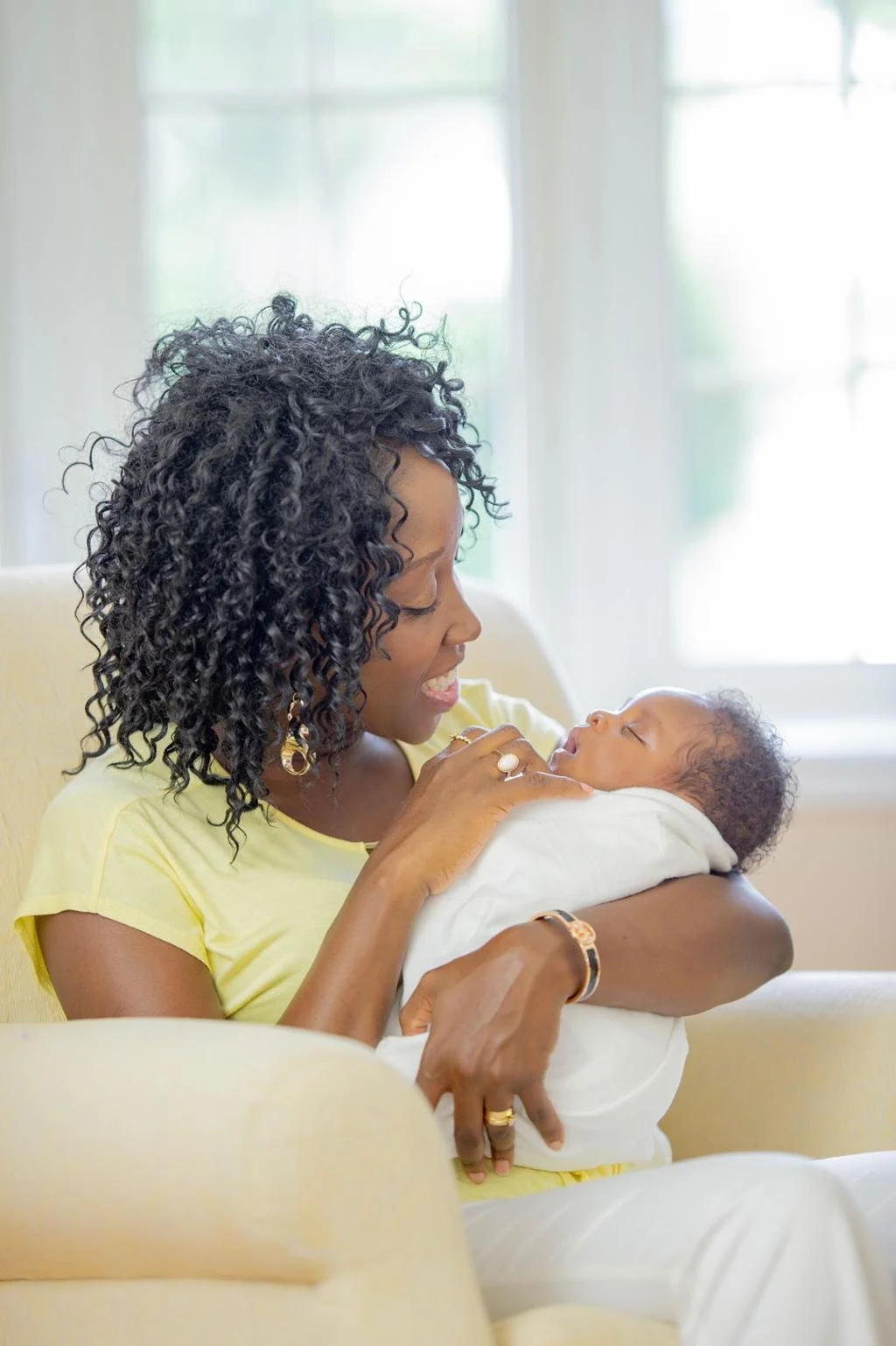 A woman with curly black hair holding a sleeping baby wrapped in white blanket.