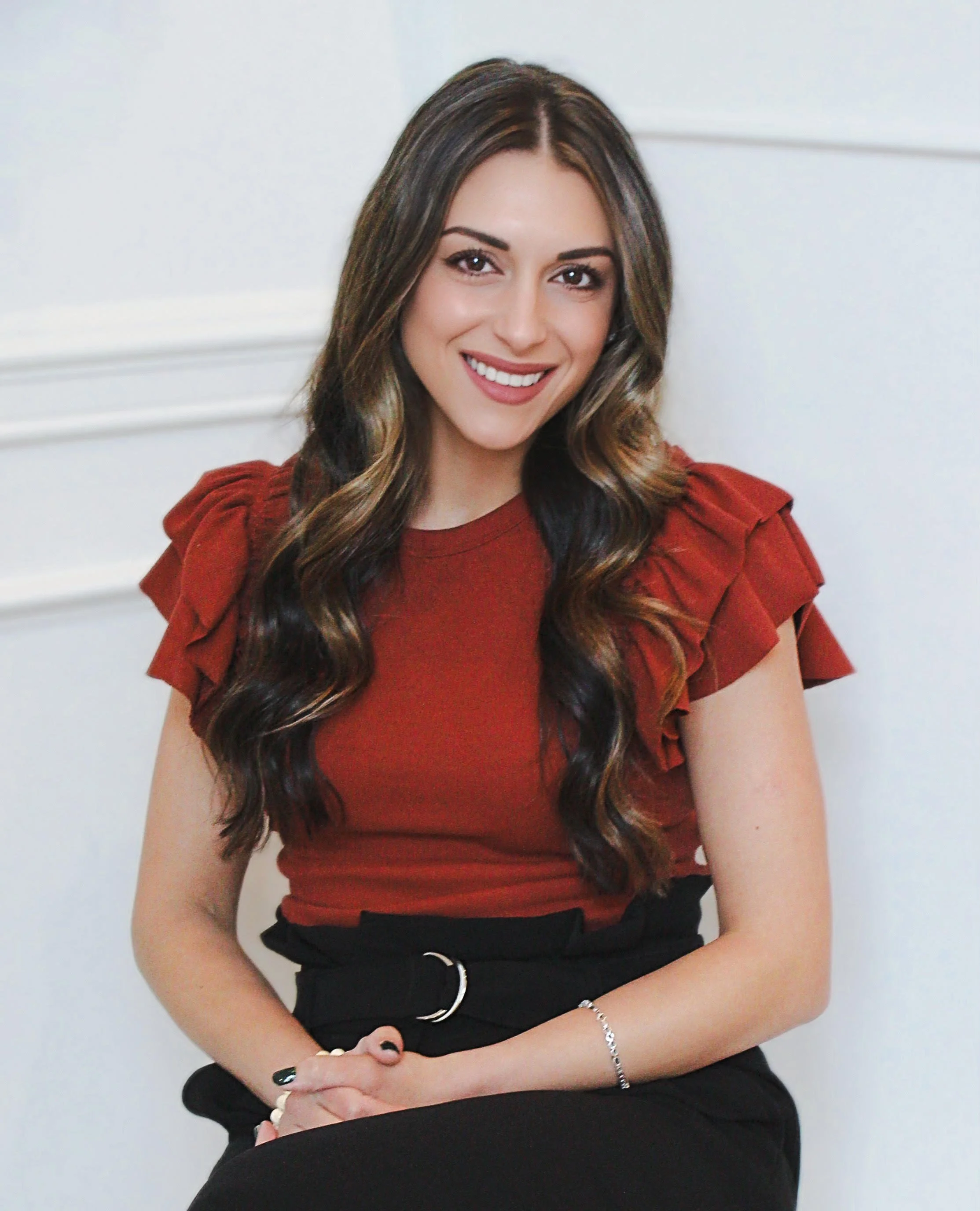 A young woman with long wavy brown hair, smiling, wearing a red top with ruffled sleeves and black pants, sitting with her hands clasped in her lap indoors.