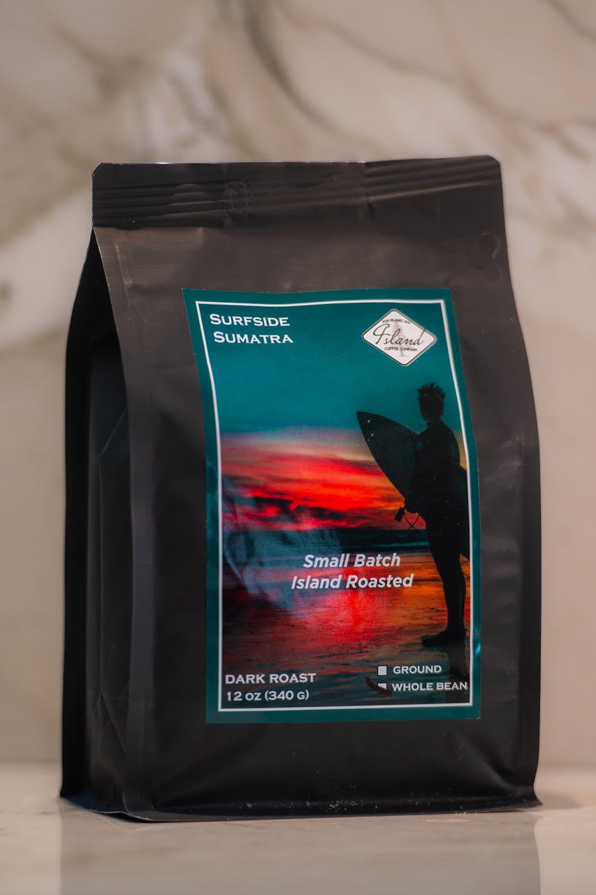 A black coffee bag labeled 'Surfside Sumatran,' featuring a sunset with a silhouette of a person holding a surfboard, indicating small batch, island roasted dark roast coffee, 12 ounces.