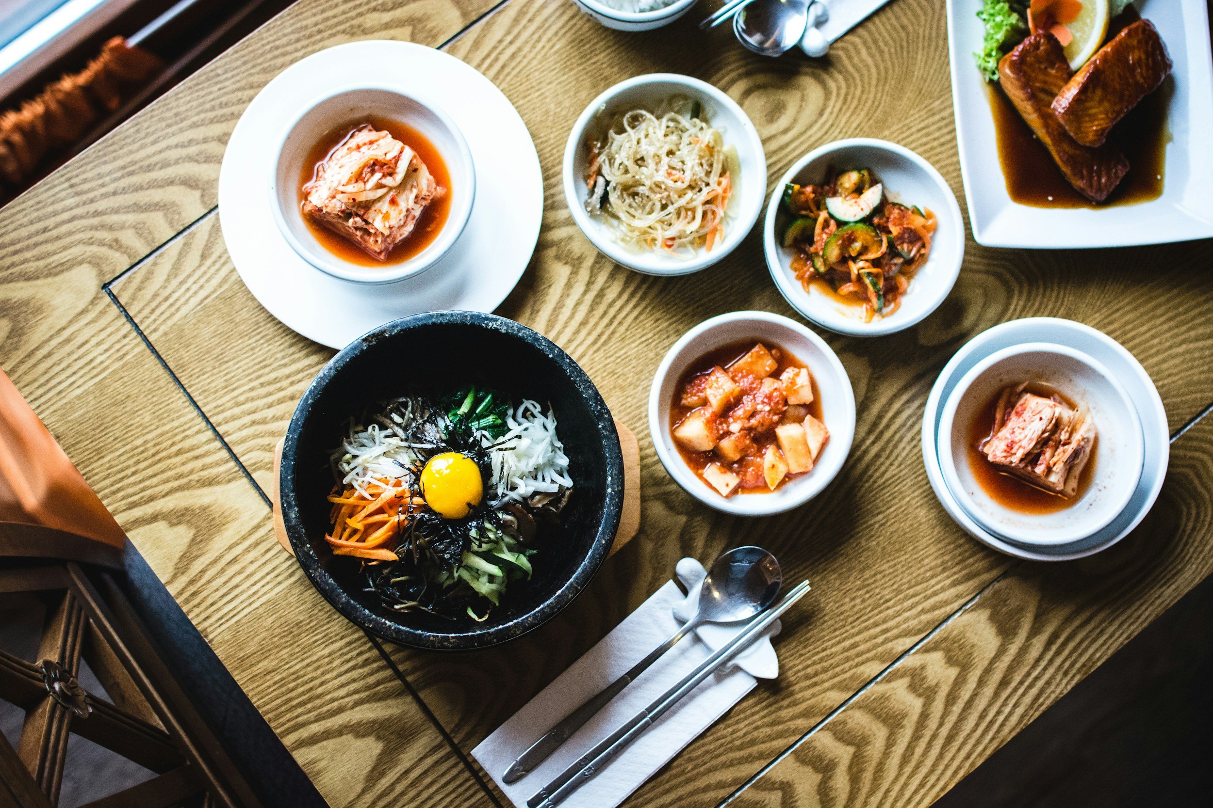 A table with various traditional Korean side dishes, including kimchi, seasoned vegetables, and rolled meat, surrounding a bowl of bibimbap topped with raw egg yolk and vegetables.