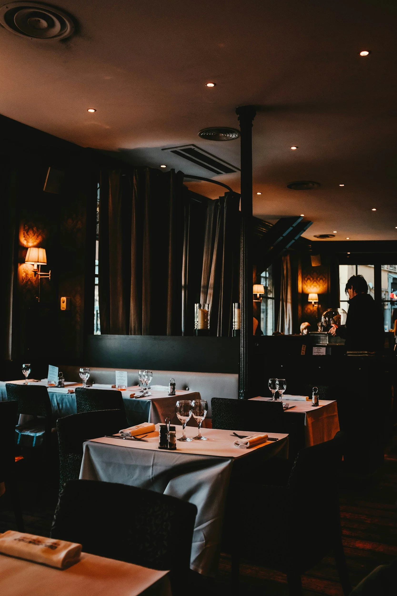 Interior of an upscale restaurant with white-clothed tables set with wine glasses, silverware, and napkins, dim lighting, dark walls, and a server in the background.