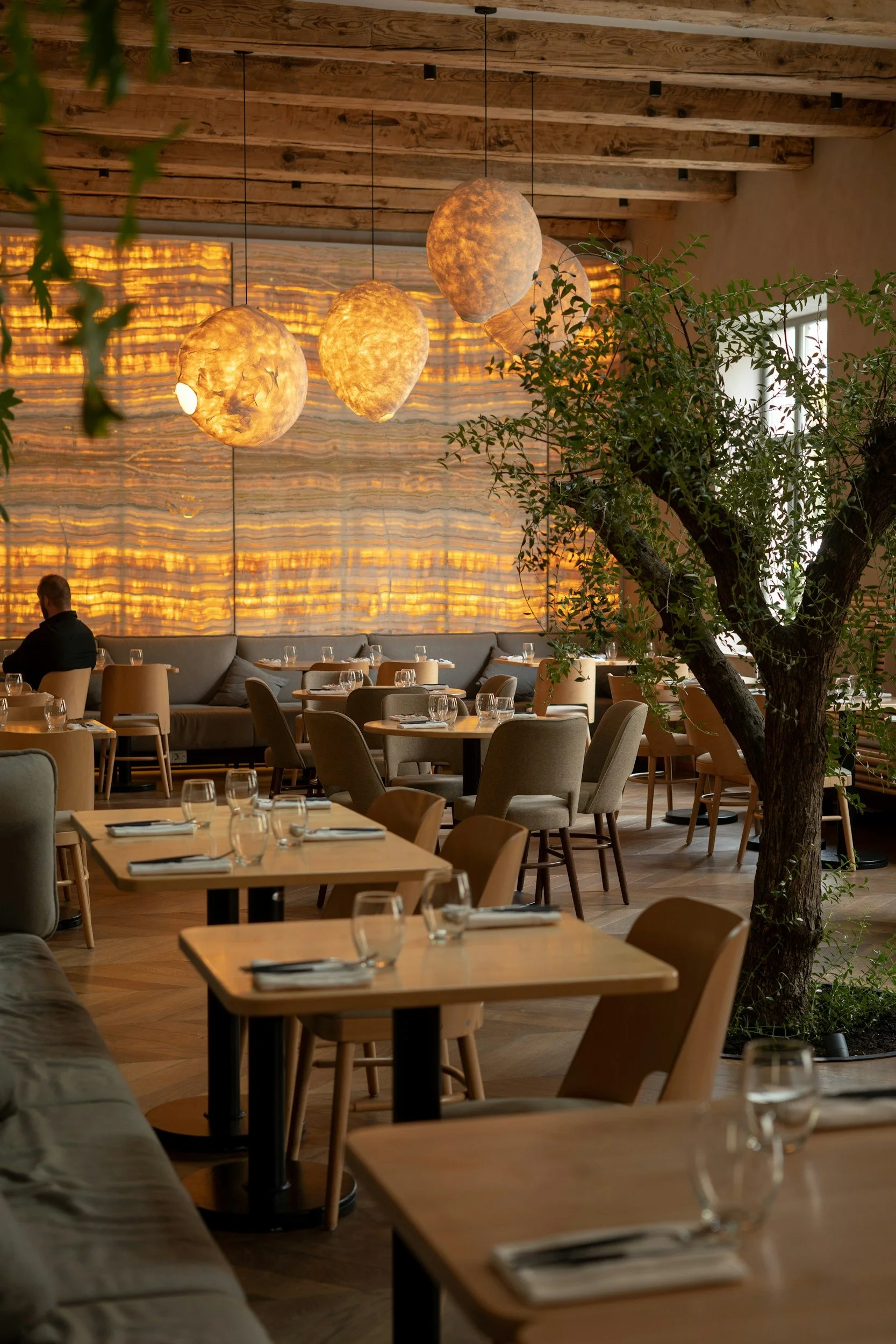 An interior view of a restaurant with wooden ceilings, hanging spherical light fixtures, and a mix of chairs and tables set with glassware and napkins. There is a large tree to the right and a person seated in the background.
