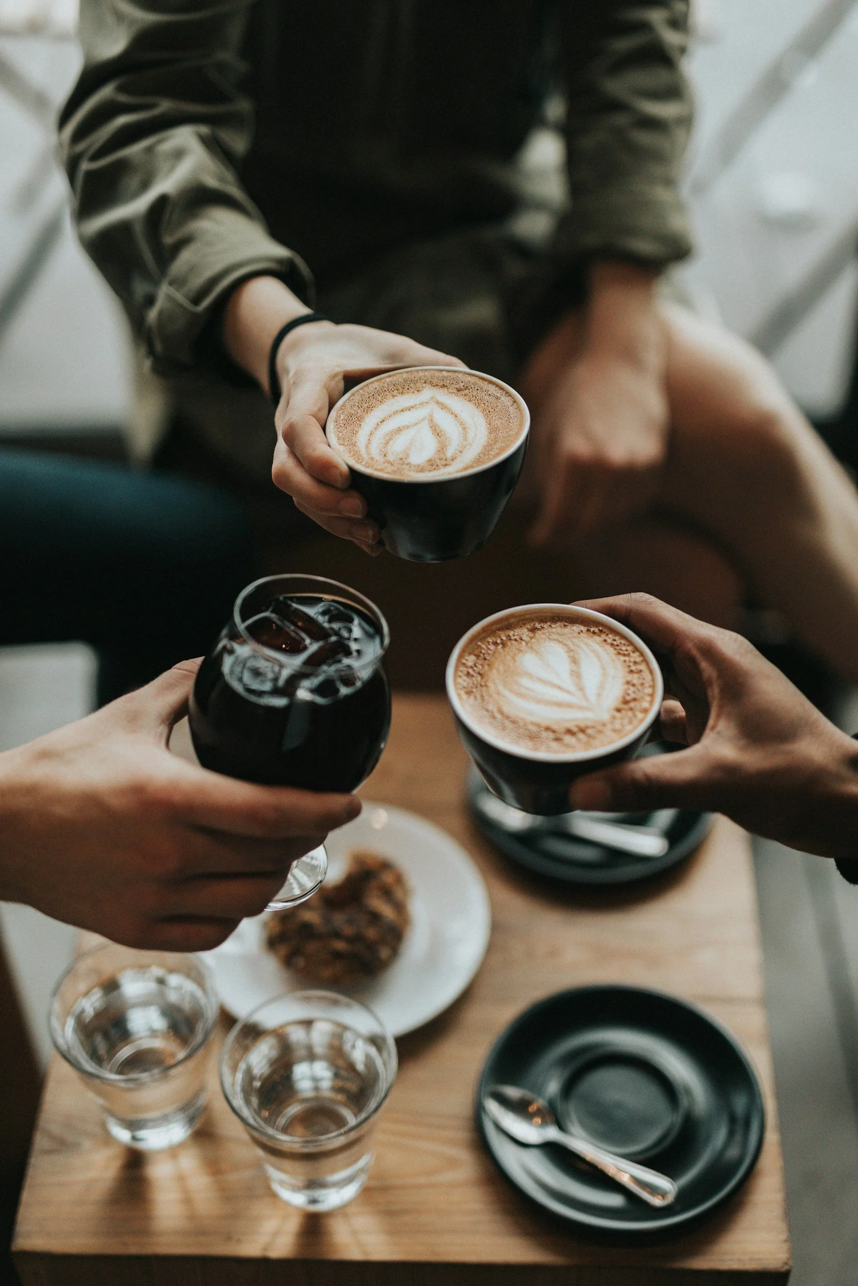 Three people are toasting with coffee and a glass of iced drink at a wooden table in a cafe, with a plate of food and empty glasses also on the table.