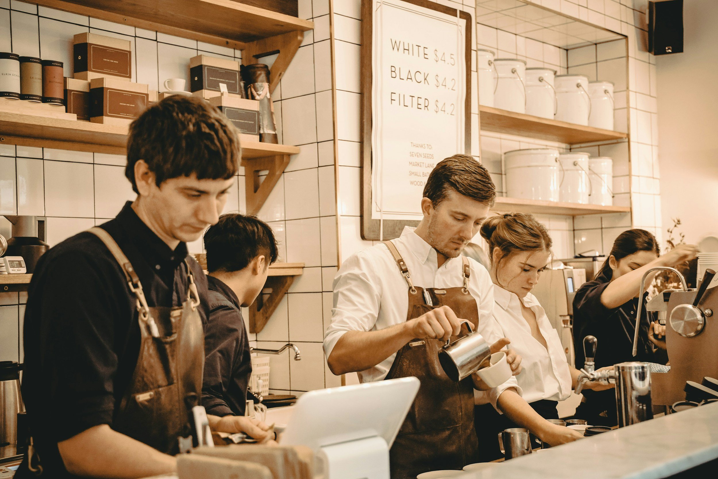 Baristas working behind the counter in a coffee shop, preparing beverages for customers, with white tiled walls, wooden shelves, and a menu board displaying prices for different types of coffee.