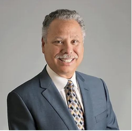 A professional-looking man with curly gray hair and a mustache, wearing a dark blue suit, white shirt, and patterned tie, smiling against a plain gray background.