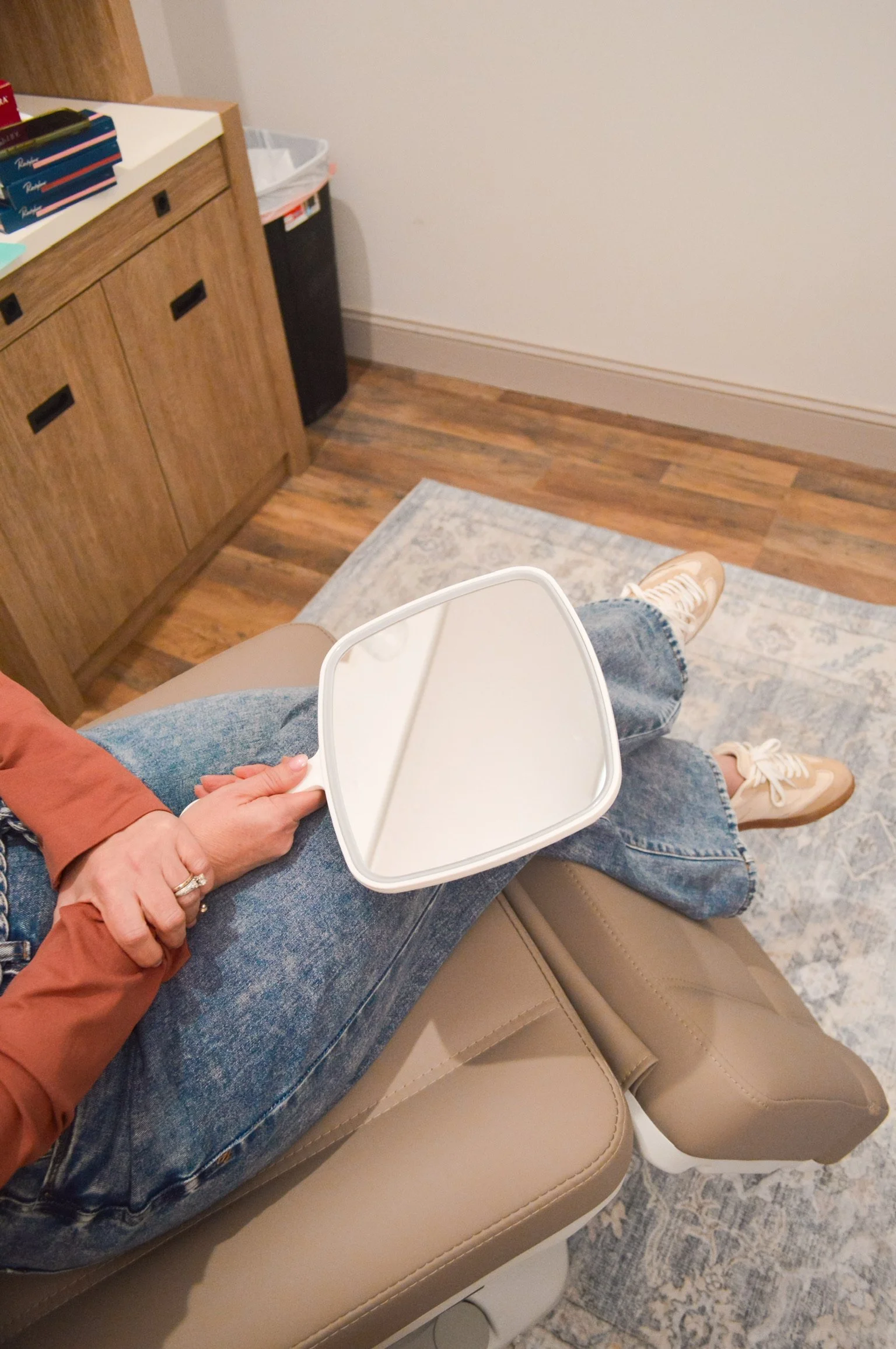 Person sitting in a medical or dental office holding a small mirror, with legs crossed, wearing denim jeans and beige sneakers, resting on a beige leather chair in a room with wood flooring, a rug, and a cabinet.