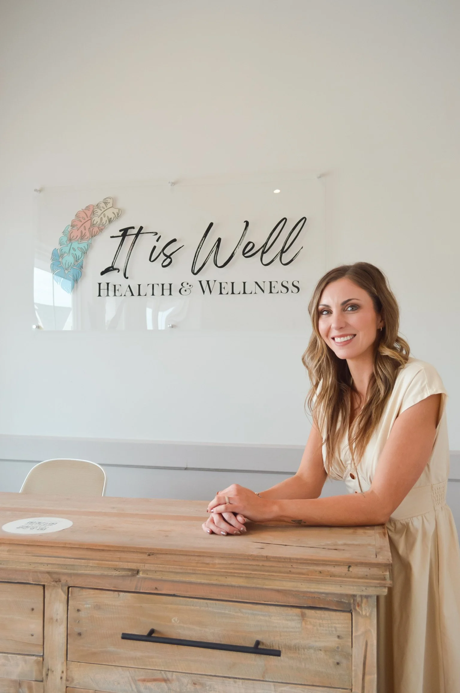 A woman sitting at a wooden desk with a sign behind her that reads 'It is Well Health & Wellness' with a stylized brain illustration in pink, blue, and beige colors.