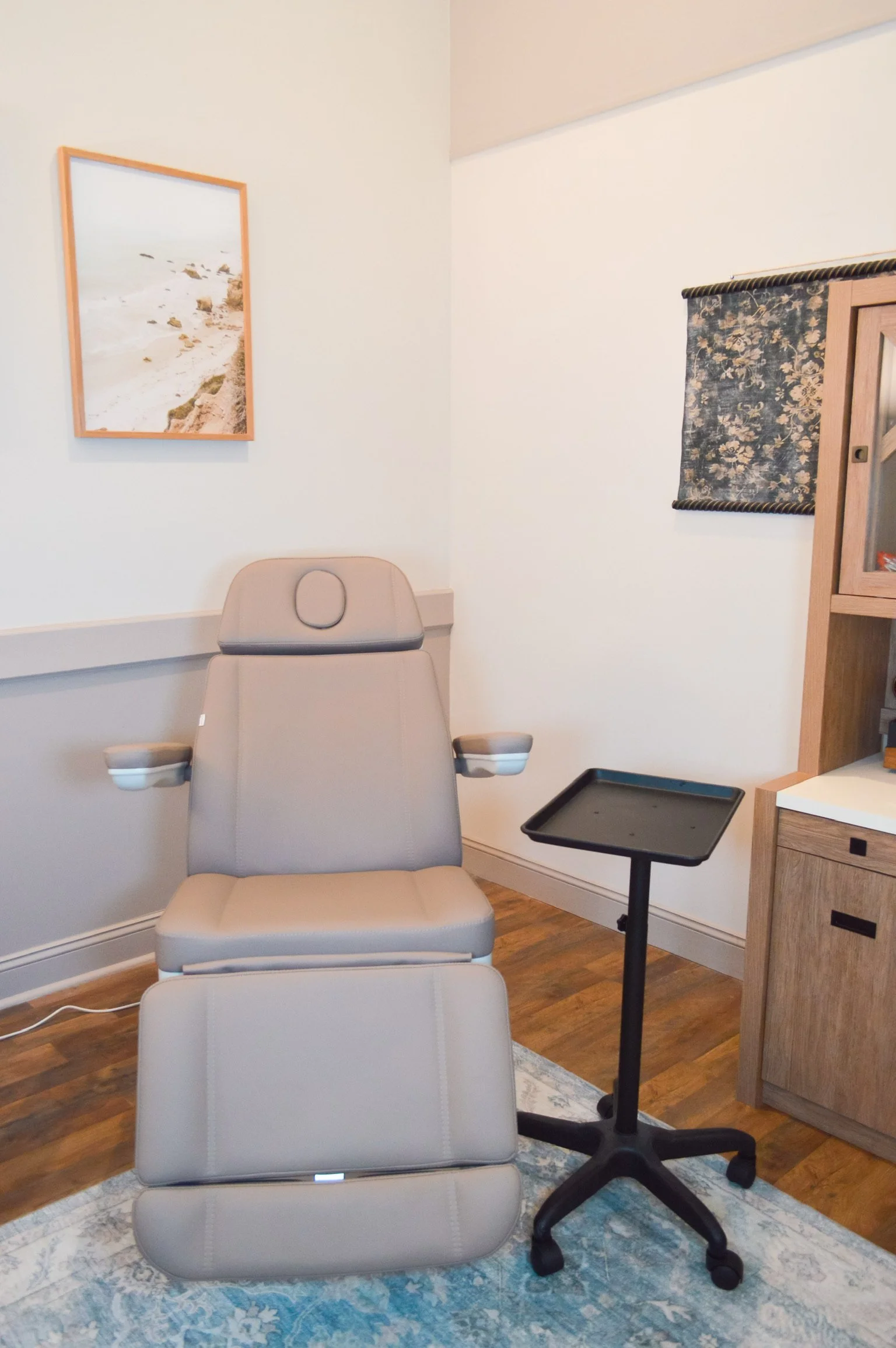 Medical examination chair in a room with hardwood floor, a small side table, a wooden cabinet, and a framed picture of a beach scene on the wall.