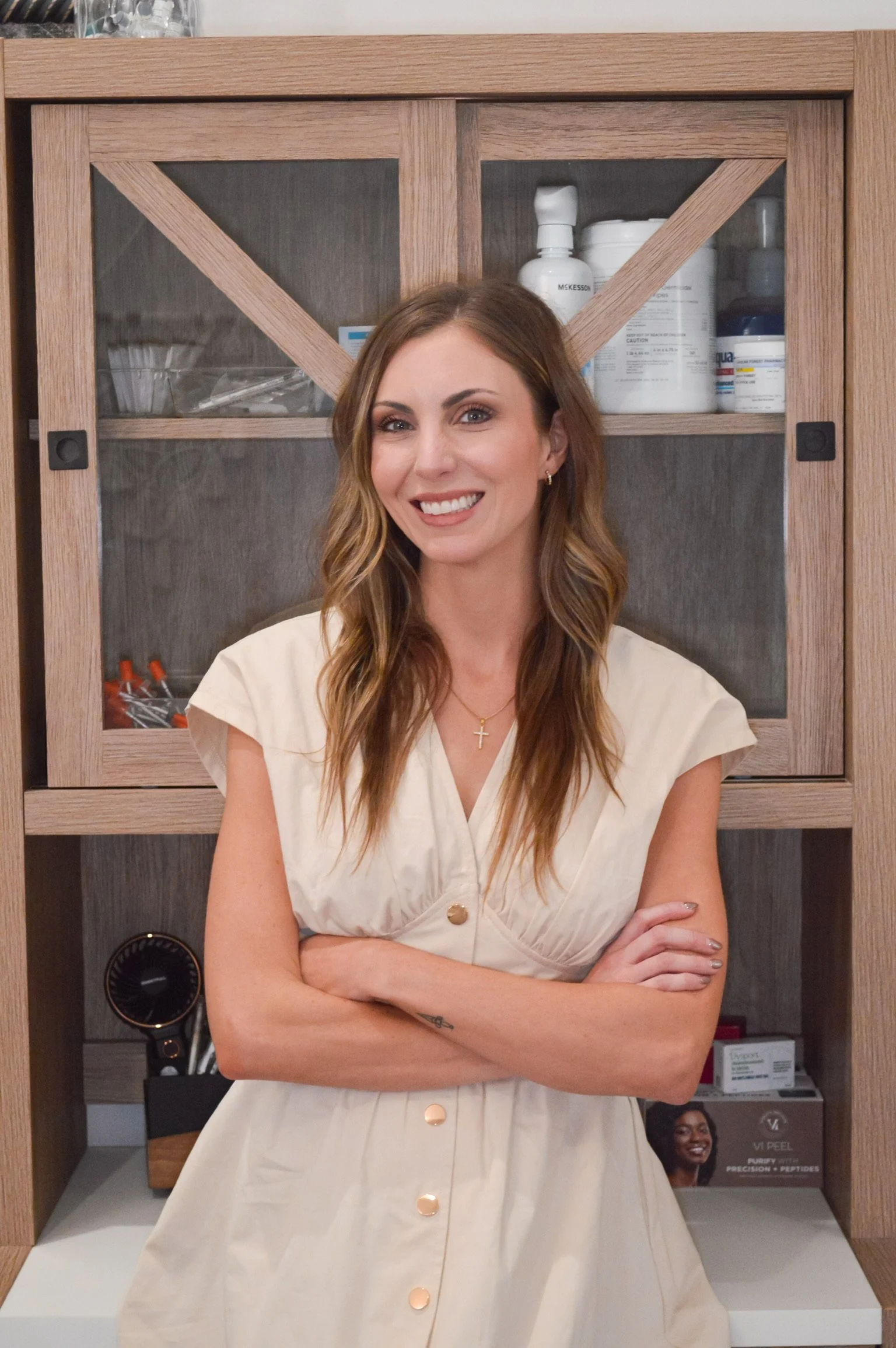 A woman with long brown hair, wearing a cream-colored dress with gold buttons, standing with arms crossed in front of a wooden cabinet with glass doors. The cabinet contains medical supplies and equipment.