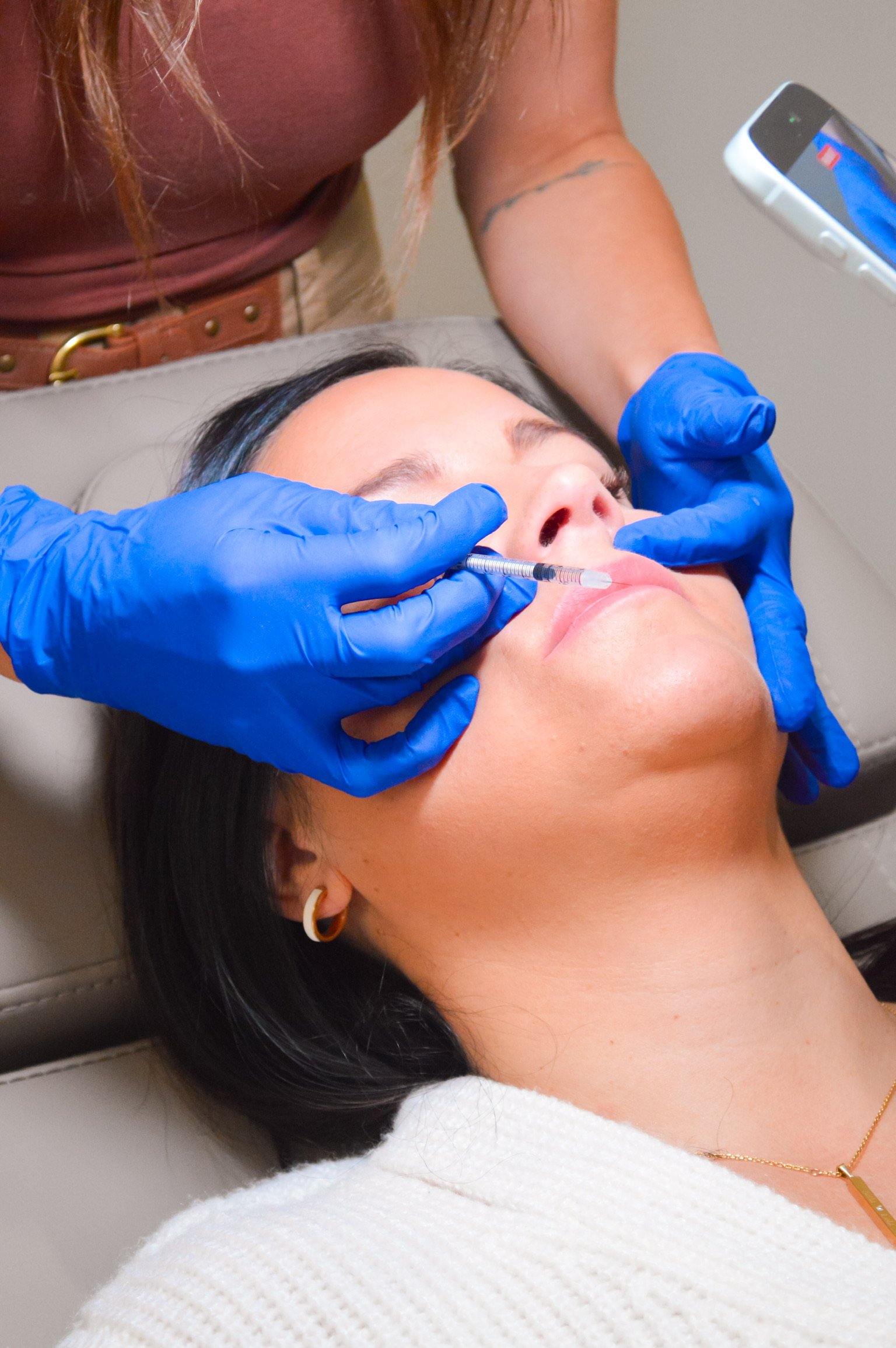 A woman receiving a cosmetic injection in her lips from a healthcare professional wearing blue gloves.