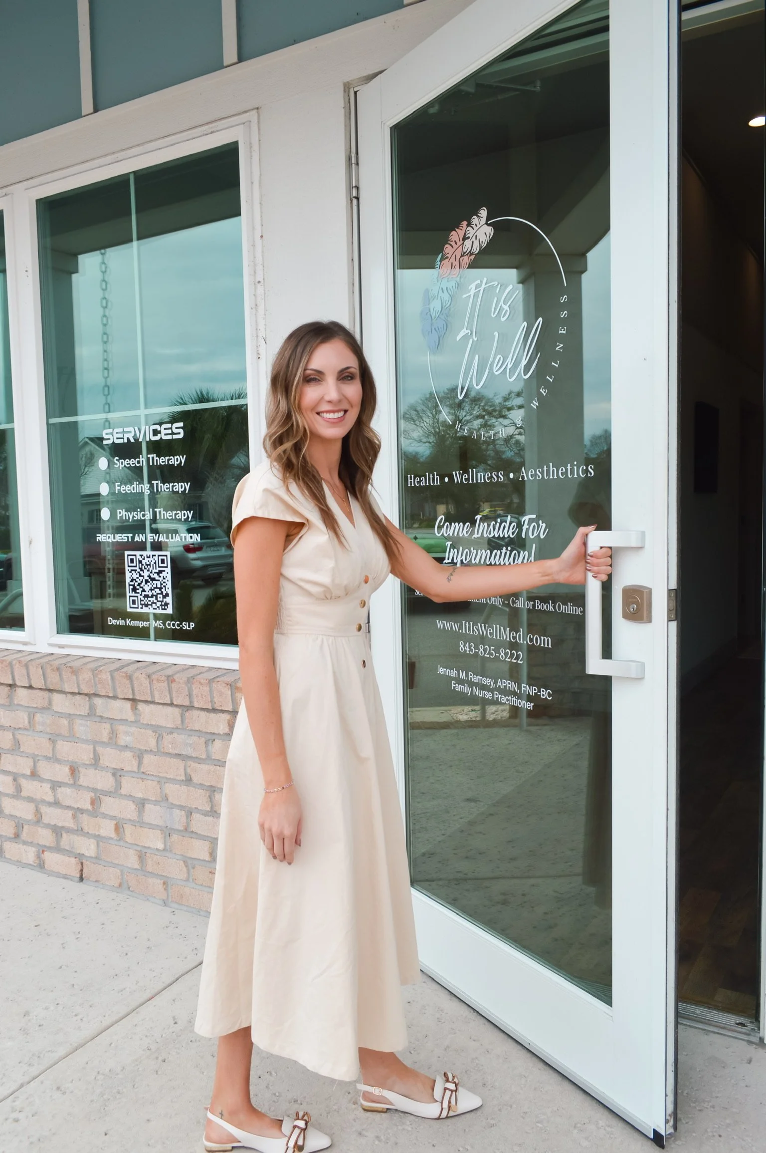 A woman with long wavy hair wearing a beige dress and white shoes holding open the door of a wellness clinic named 'It's Well' with the text 'Health, Wellness, Aesthetics'. The clinic's window displays services including speech therapy, feeding therapy, and physical therapy.