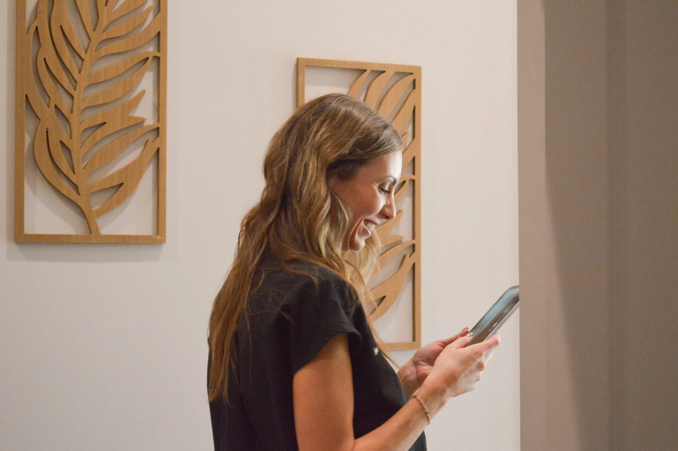 A woman with long wavy hair smiling while looking at her smartphone in an indoor setting with wooden decorative wall art.