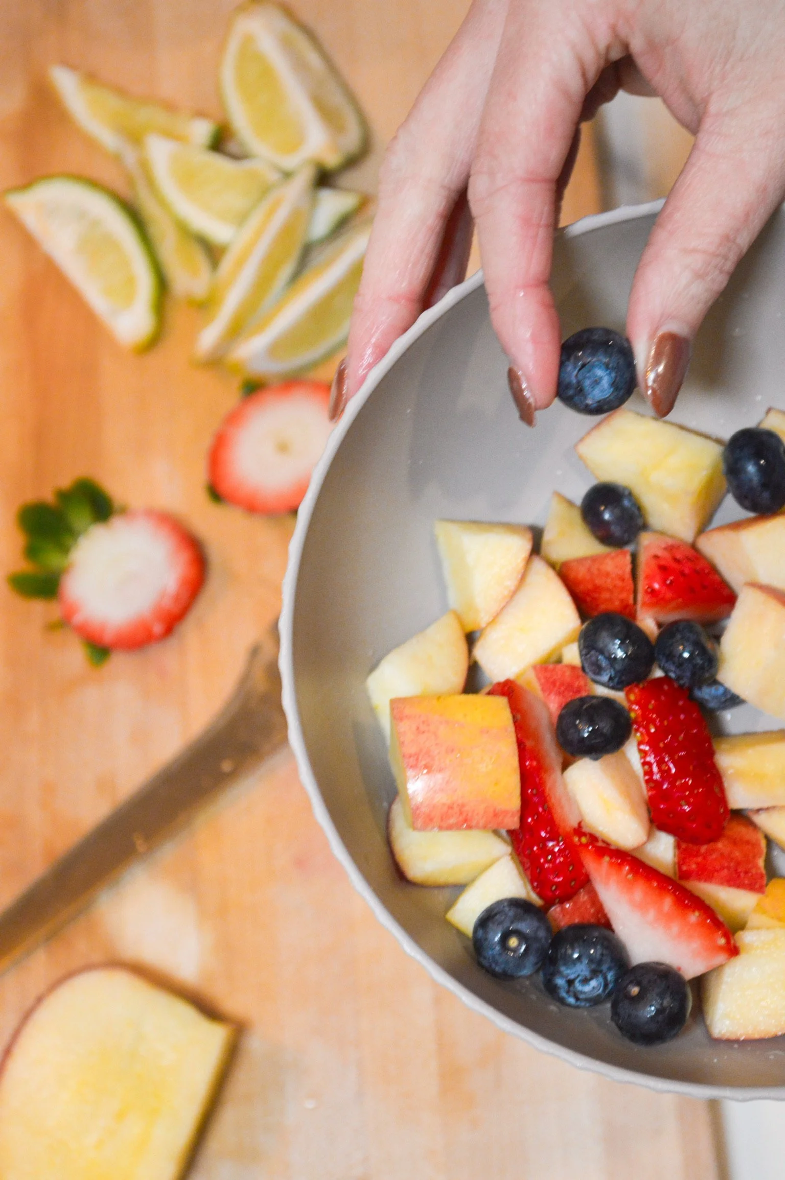 A hand holding a bowl of cut-up mixed fruit including strawberries, blueberries, and apples. In the background, sliced lemons and strawberries on a cutting board, along with a knife.