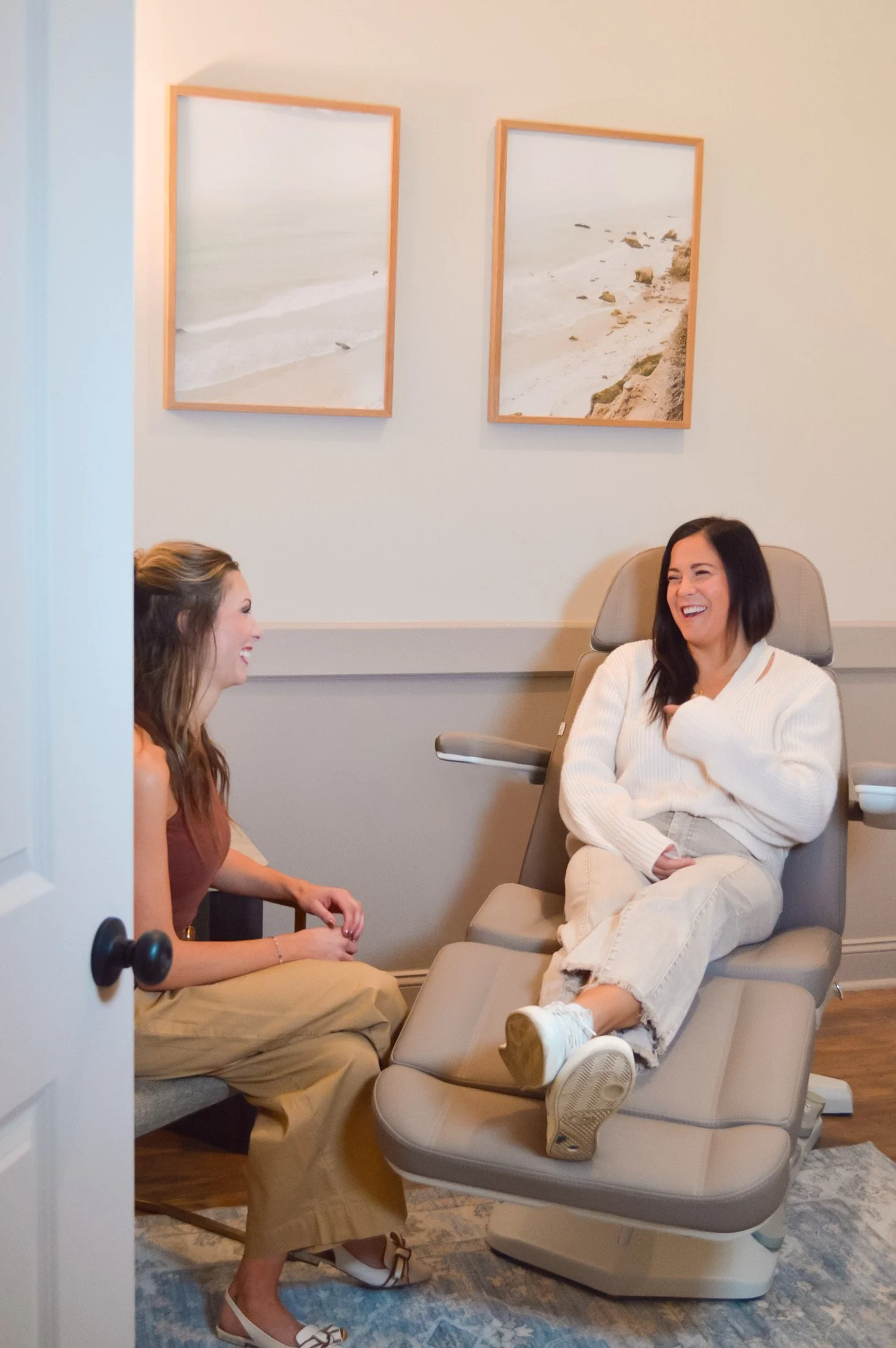 Two women laughing together in a medical office, one seated on an examination chair and the other on a sofa, with beach pictures on the wall behind them.