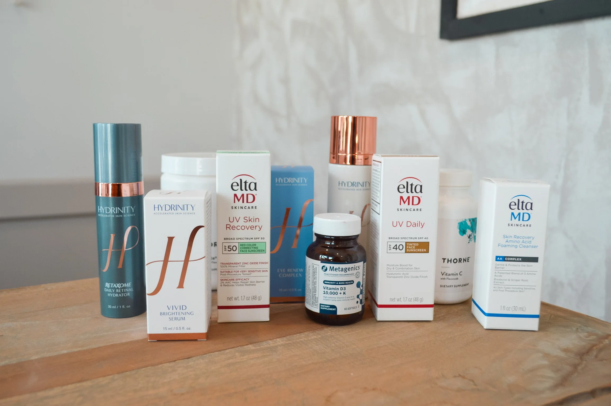 Collection of skincare and supplement products on a wooden surface, including EltaMD, Hydrinity, Thorne, and Metagenics brands with various lotions, serums, sunscreens, and vitamins.