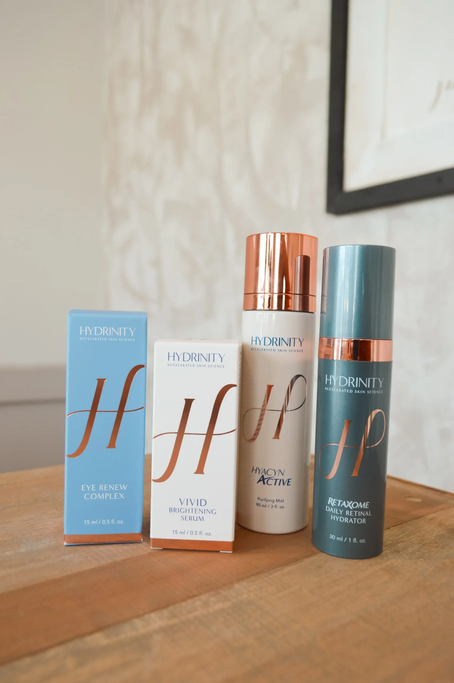 Four skincare products from the brand Hydrinity arranged on a wooden surface. The products include Eye Renew Complex, Vivid Brightening Serum, Hyacin Active Purifying Mist, and Retaxome Daily Retinal Hydrator, with packaging in blue, white, and teal with metallic accents.