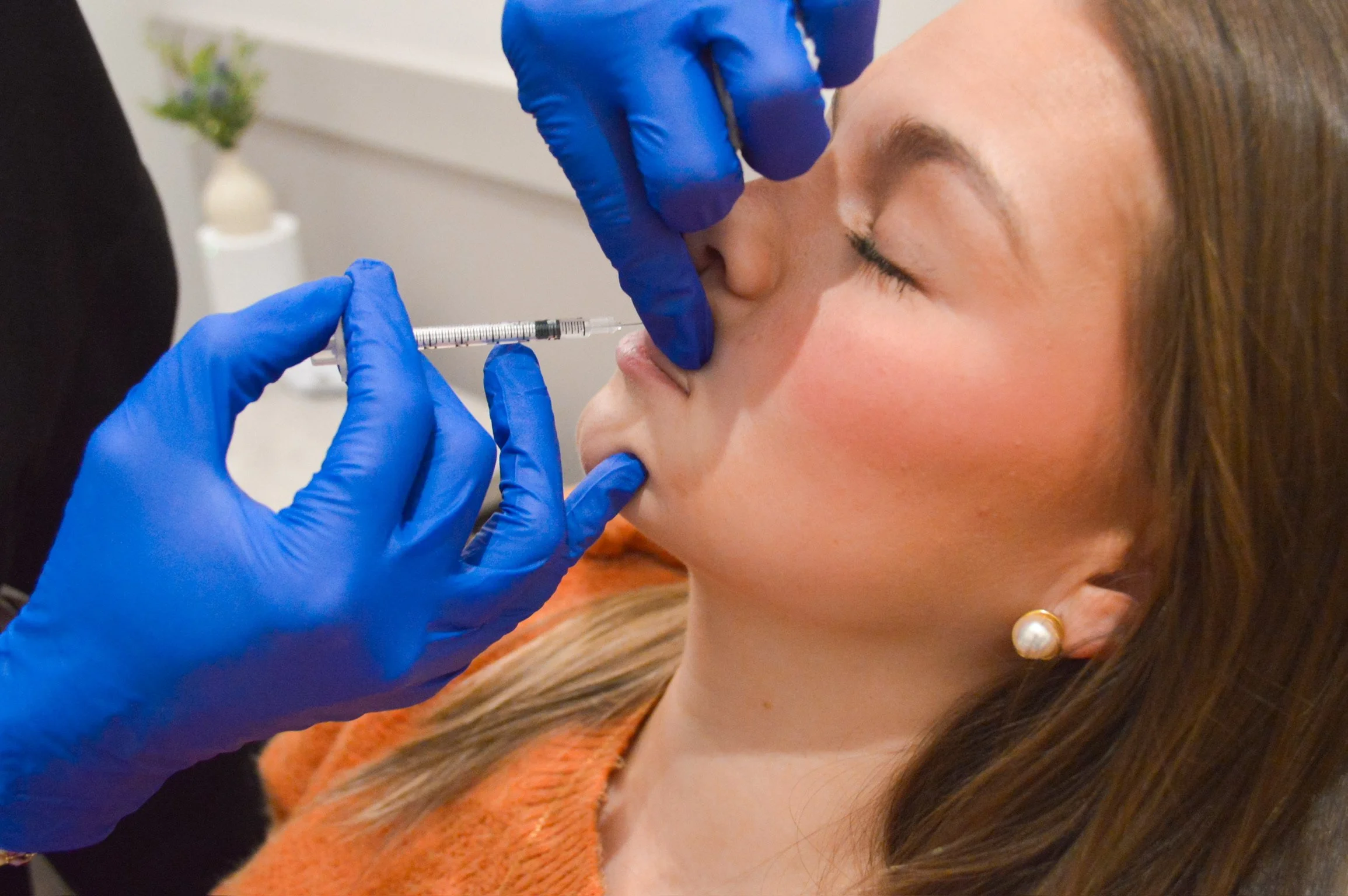 A woman receives a cosmetic injection in her nose administered by a healthcare professional wearing blue gloves.