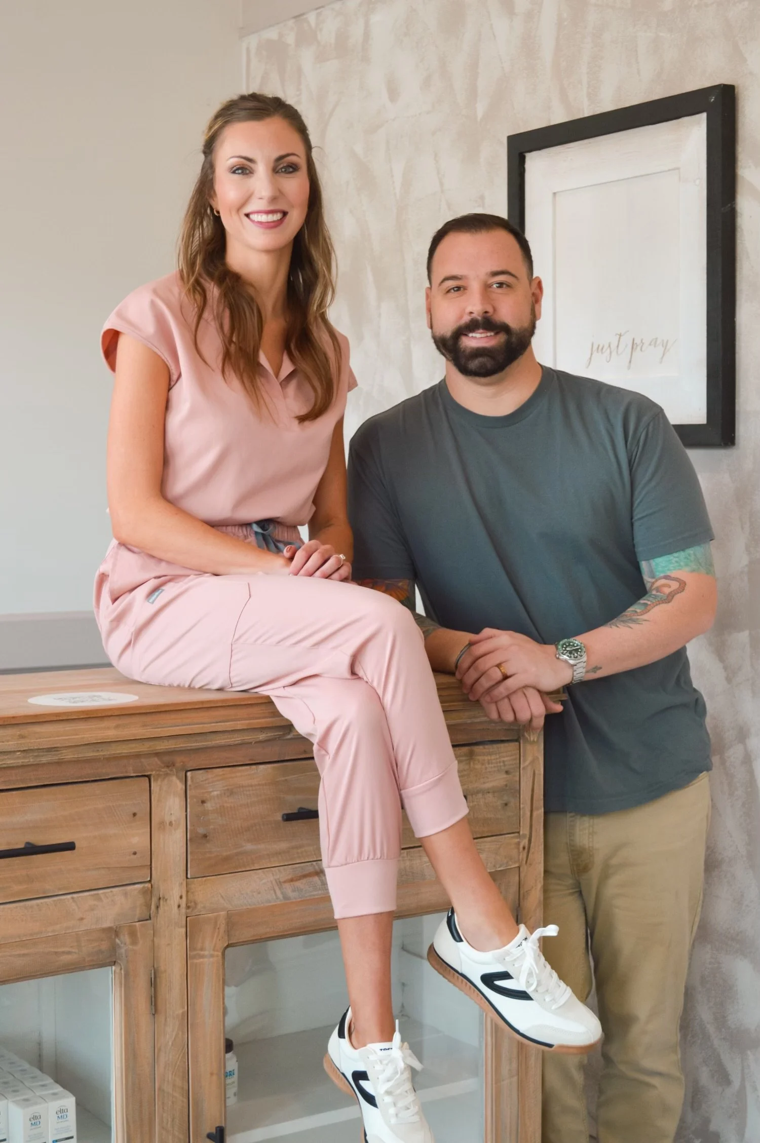 A woman sitting on a wooden cabinet next to a man standing beside her, both smiling. There is a framed art piece on the wall behind them.