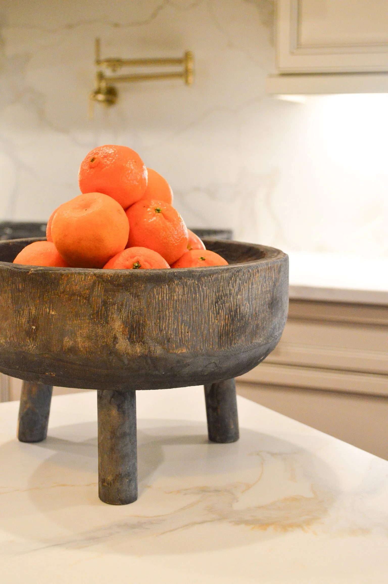 A rustic black wooden bowl filled with ripe oranges sits on a white marble surface in a kitchen.