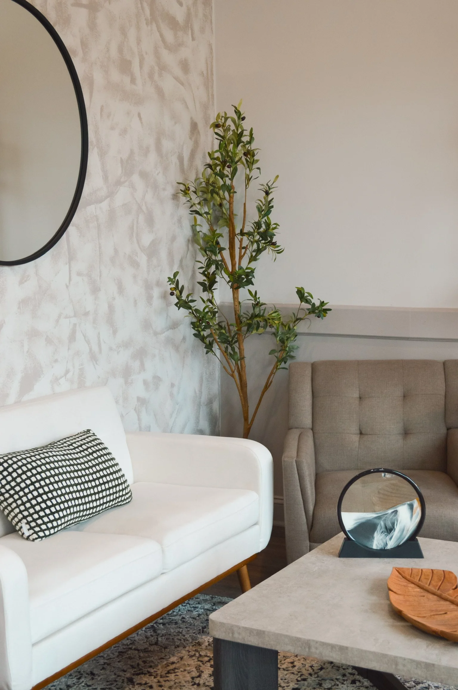 Living room with a white sofa, a beige armchair, a tall green plant, a round mirror, a coffee table with decor, and a textured wall.