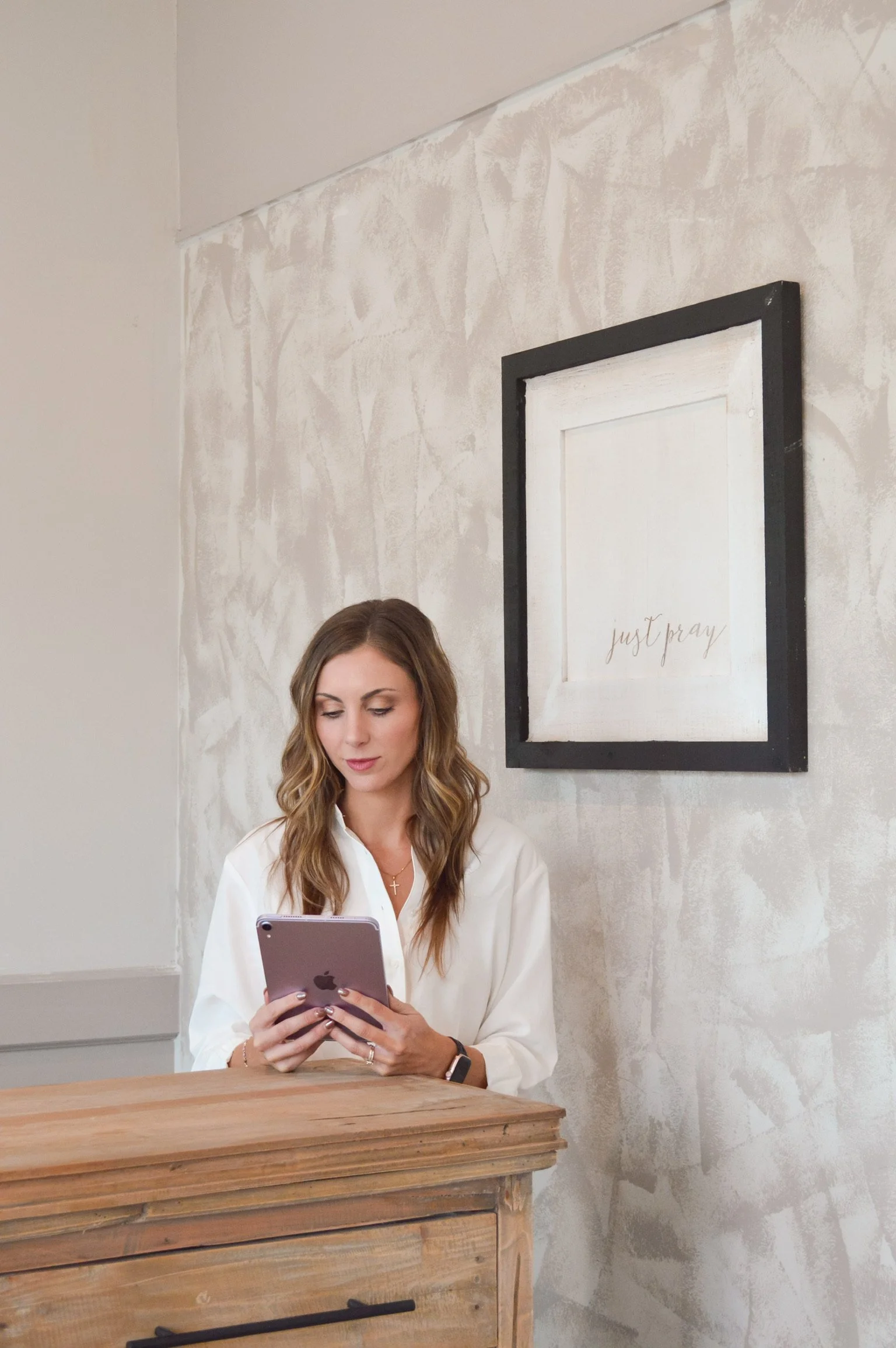 A woman with long, wavy hair wearing a white shirt, sitting behind a wooden counter, looking at her phone, with a wall and framed artwork behind her that says 'just pray'.
