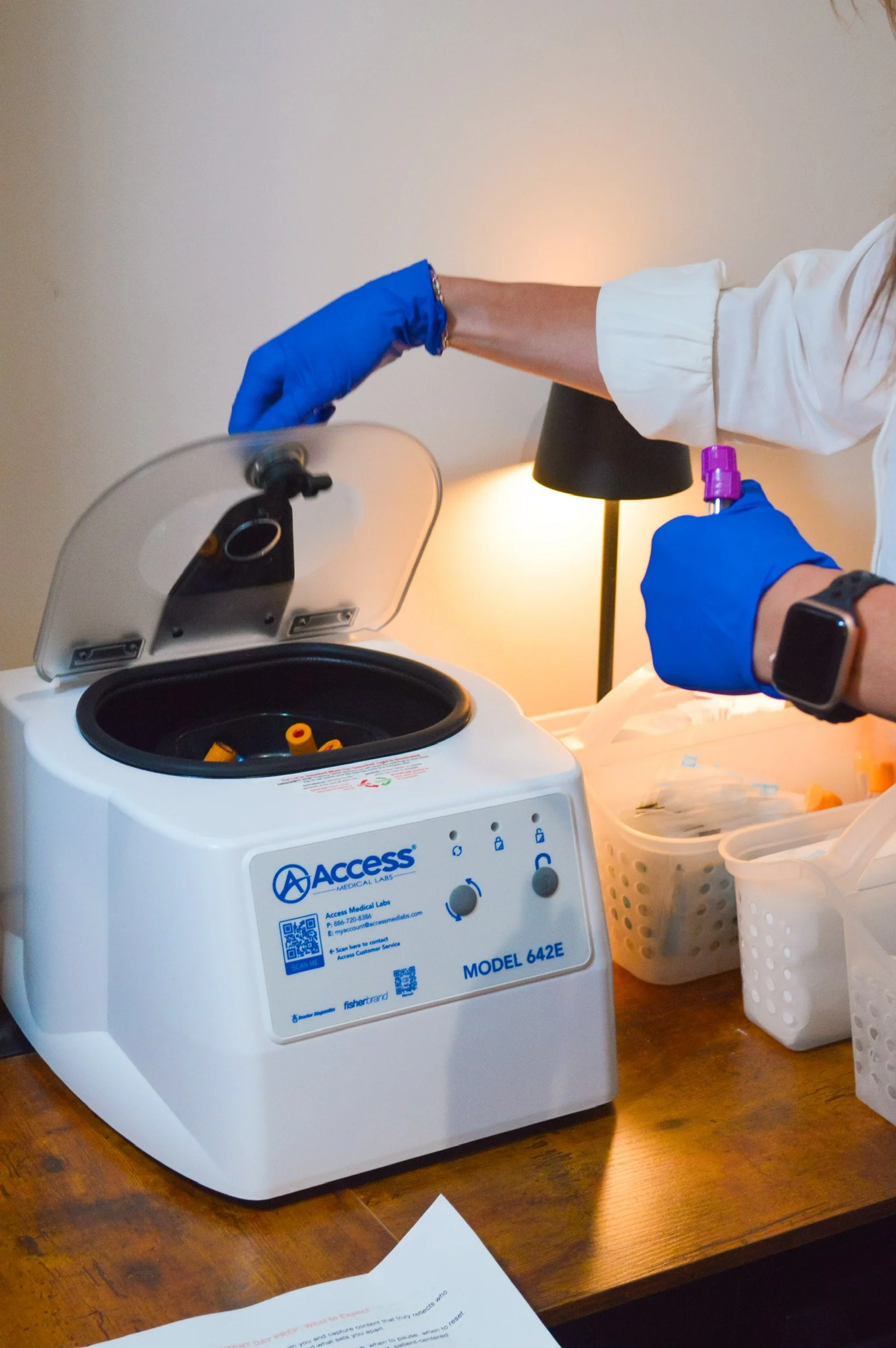A medical professional handling a centrifuge machine in a clinical setting, wearing blue gloves and a white coat.