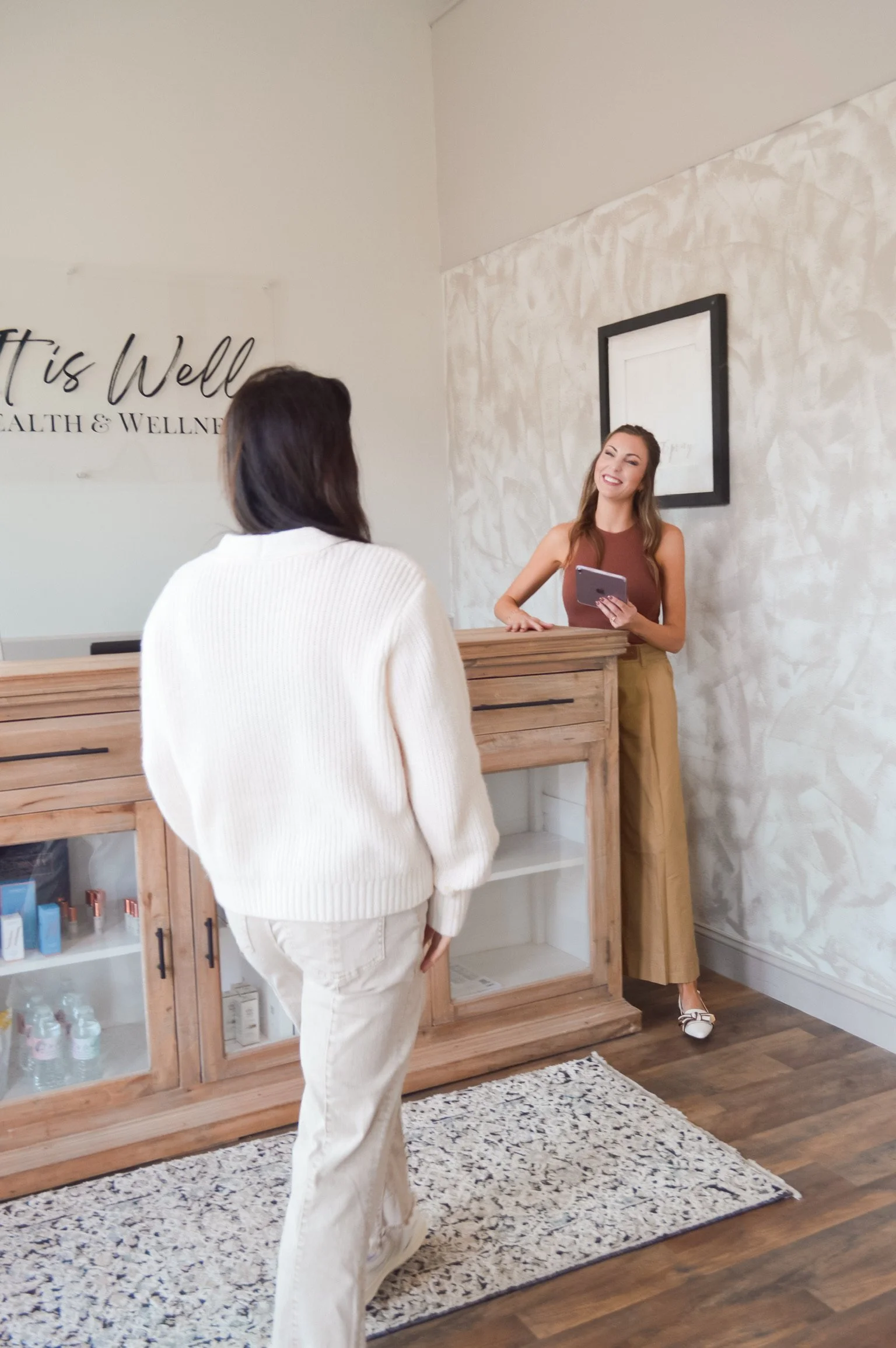 A woman working at a reception desk talking to a visitor in a wellness center or spa, with a sign on the wall that says 'It is Well' and a framed picture behind her.