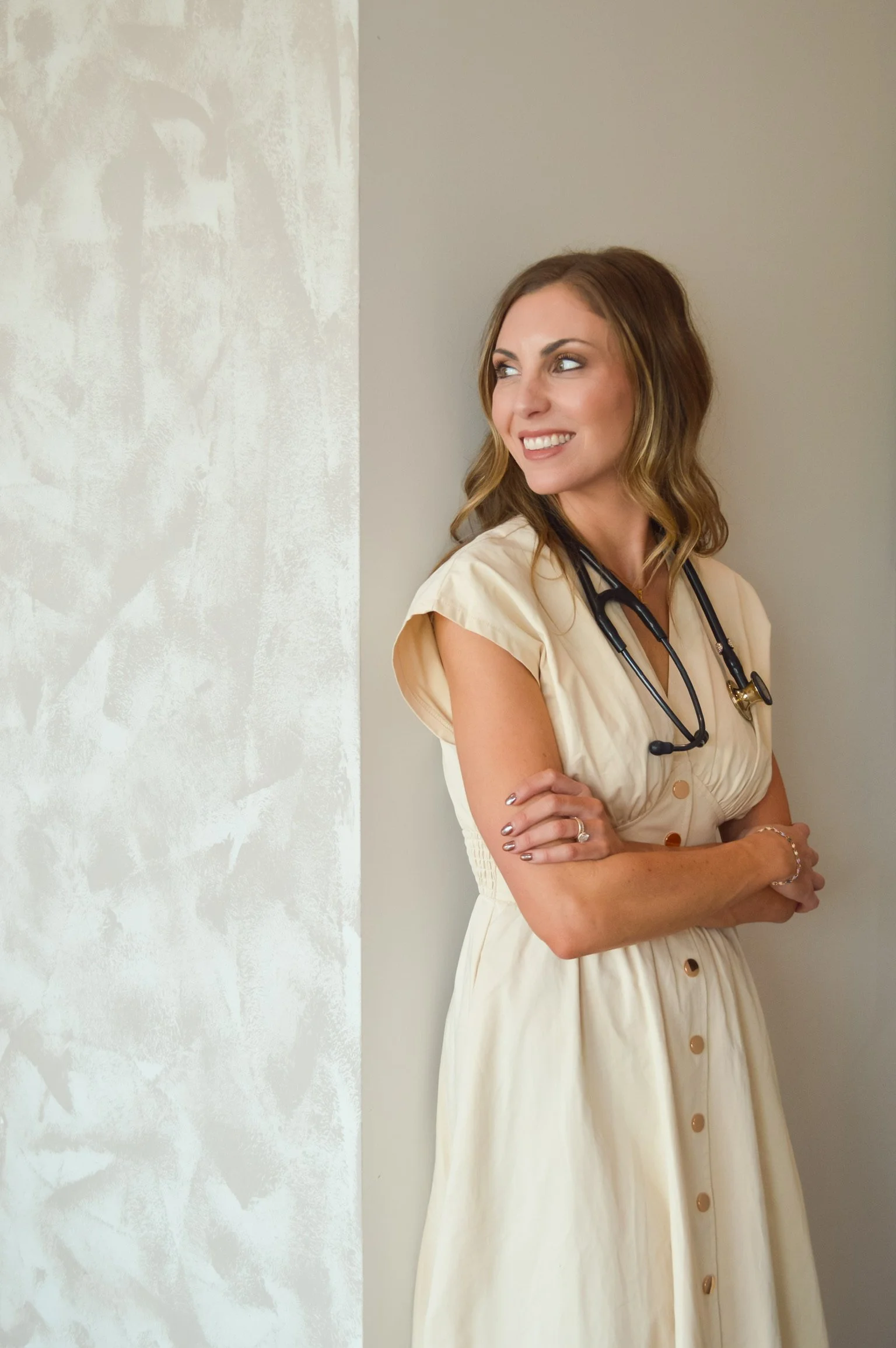 A female healthcare worker standing against a wall, wearing a beige dress with gold buttons and a stethoscope around her neck, smiling and looking off to the side.