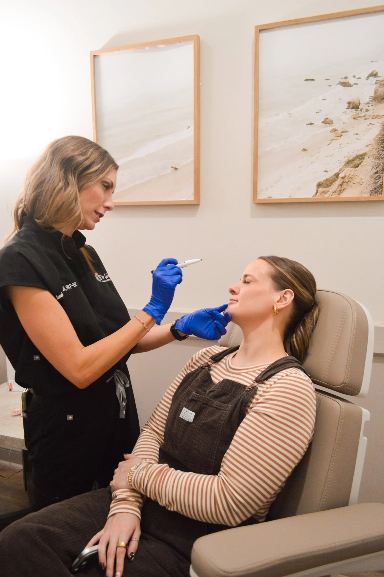 A woman is sitting in a medical chair, receiving a cosmetic injection from a healthcare professional wearing blue gloves, in a room with beach-themed artwork.