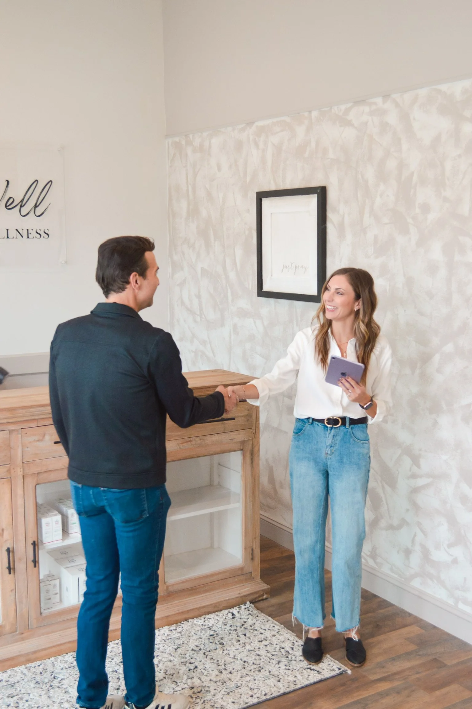 A woman and a man shaking hands in a wellness office, with the woman holding a tablet.