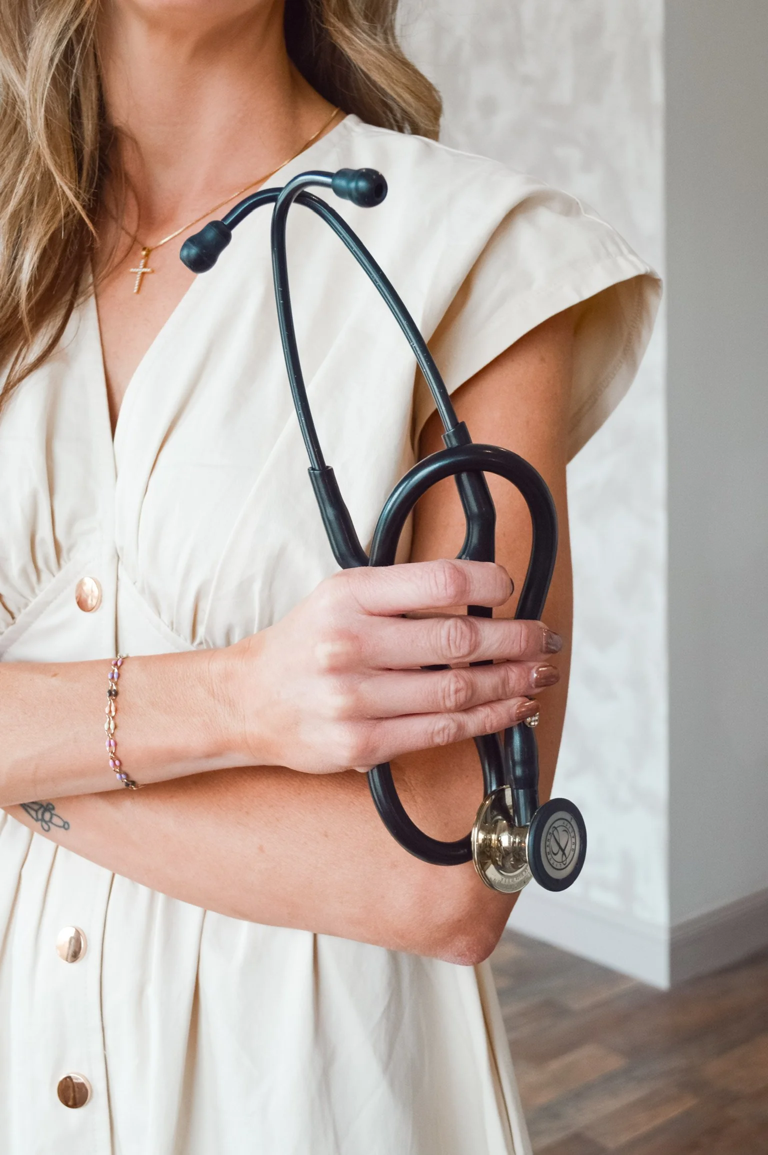 A woman holding a stethoscope on her arm, wearing a cream-colored dress with gold buttons, a delicate necklace with a cross pendant, and a bracelet.