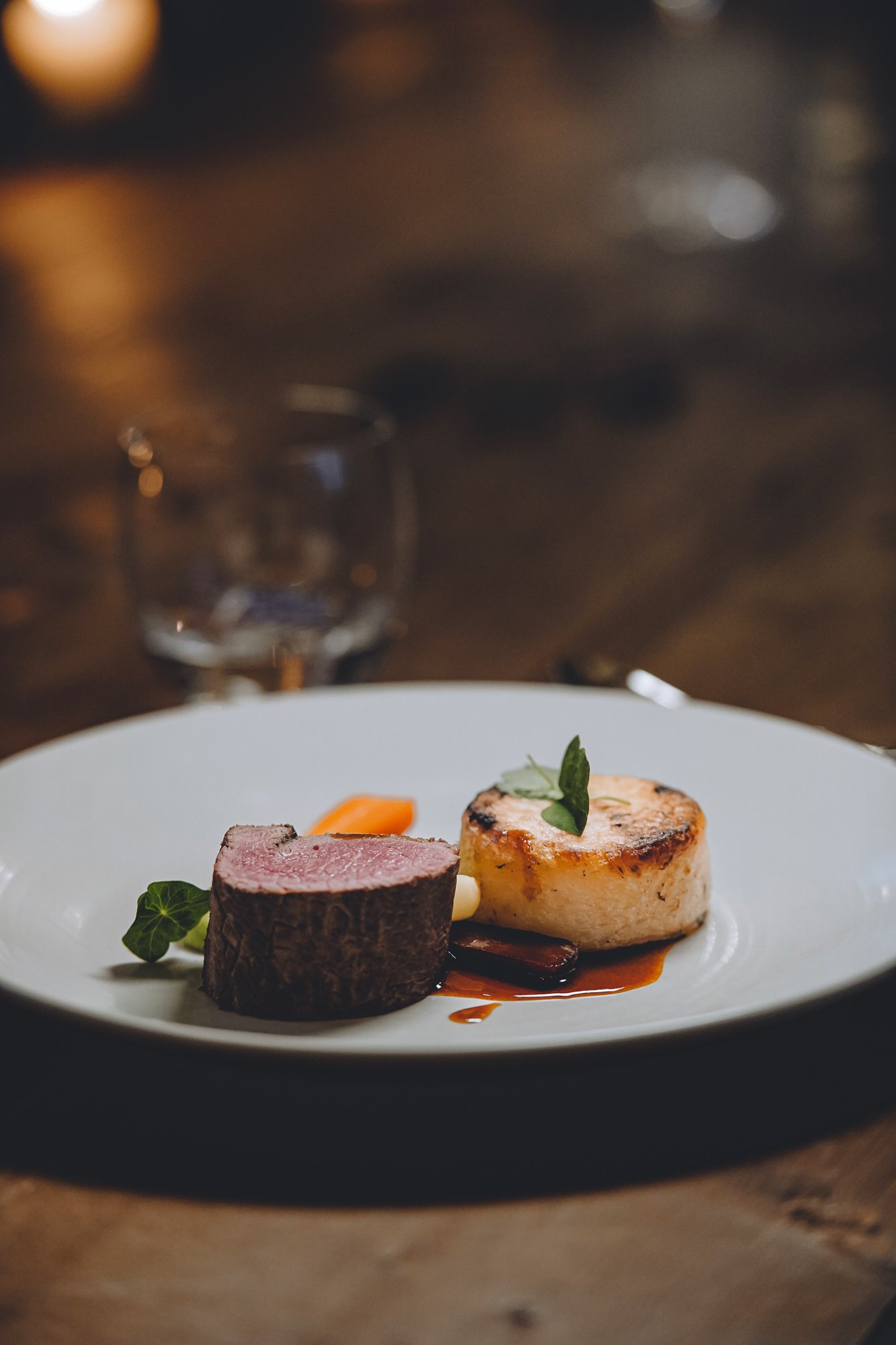 A plated gourmet meal featuring a cut of medium-rare steak and a seared scallop garnished with herbs, on a white plate with a dark background.
