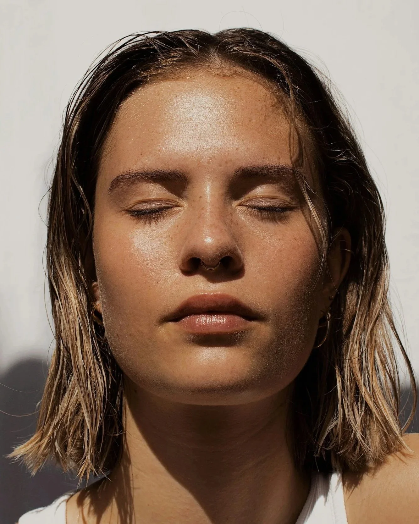 A woman with light brown hair and a septum piercing, with her eyes closed, standing against a white background.