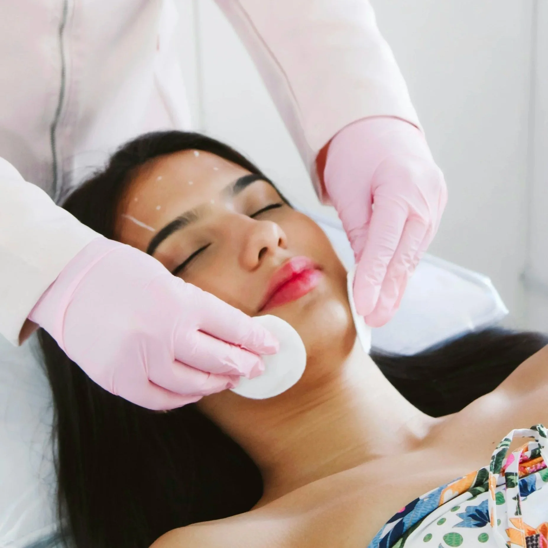 A woman with long dark hair lying on a medical bed with her eyes closed, receiving a facial treatment from a healthcare professional wearing pink gloves.