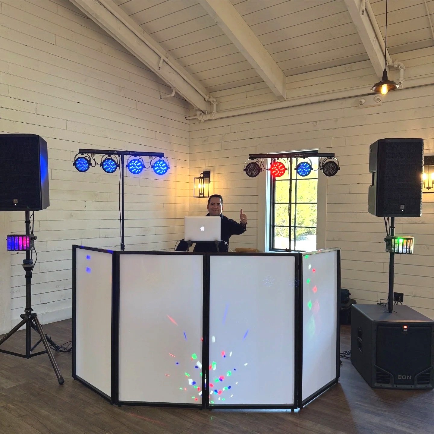 DJ man standing behind turntables with LED lighting, in a room with white painted wooden walls, speakers, and disco lights, giving a thumbs-up gesture.